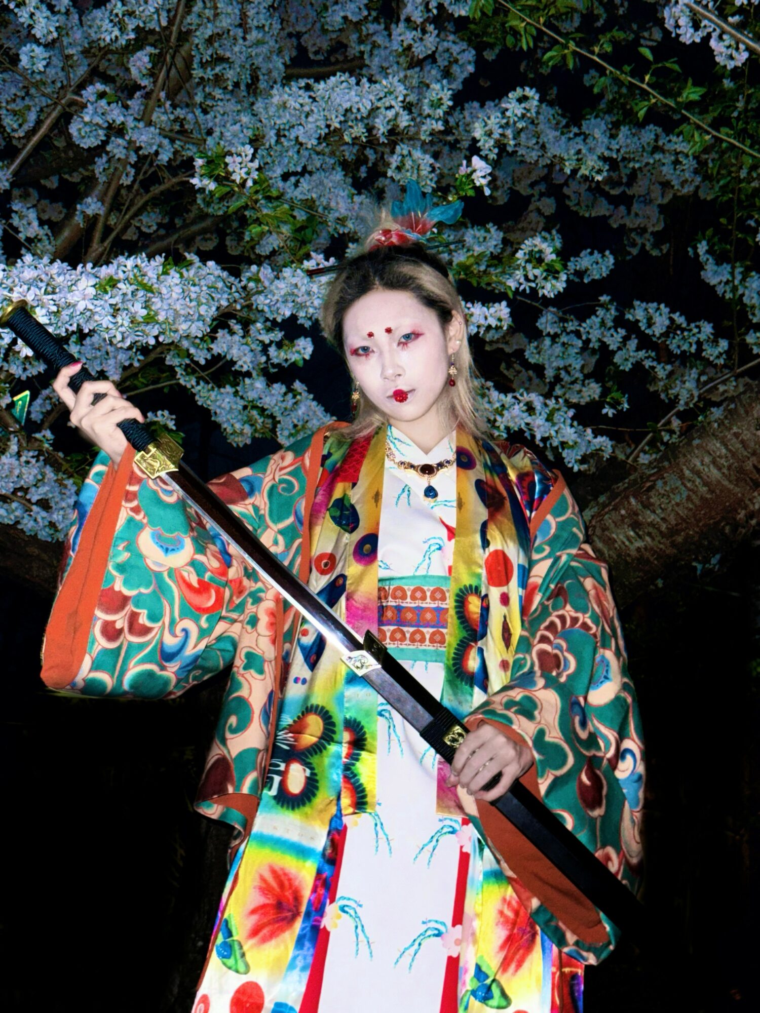 Person in colorful traditional costume holding a sword, standing in front of flowering tree at night.