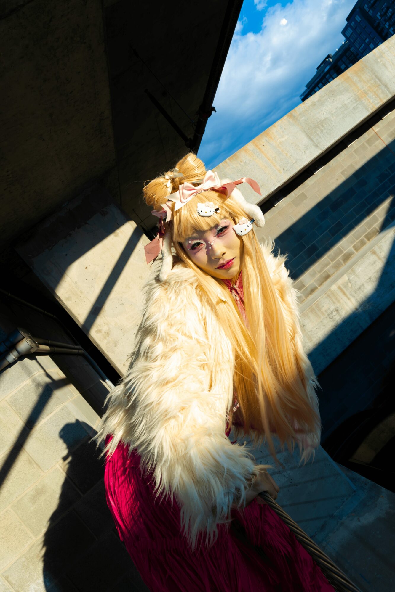 Person in elaborate costume with blonde hair, furry coat, and headpiece, standing outdoors with shadows and blue sky.