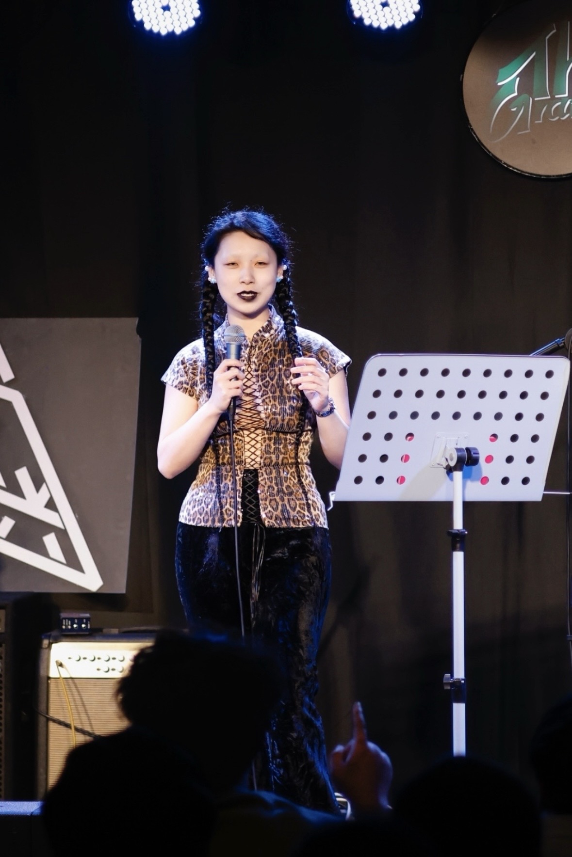 Young girl with braided hair singing on stage, holding a microphone, with a music stand nearby.