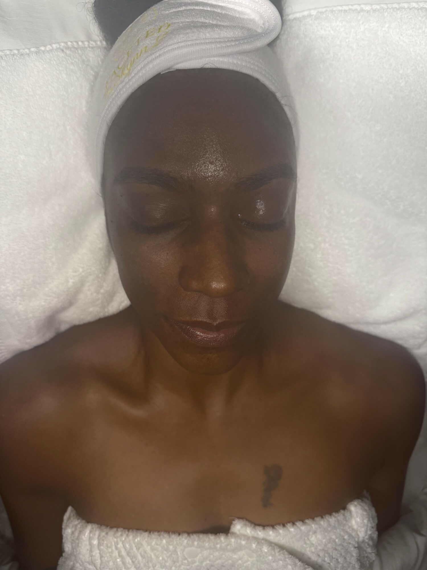 Woman with eyes closed, towel wrapped around head, lying on white surface, bare shoulders visible.