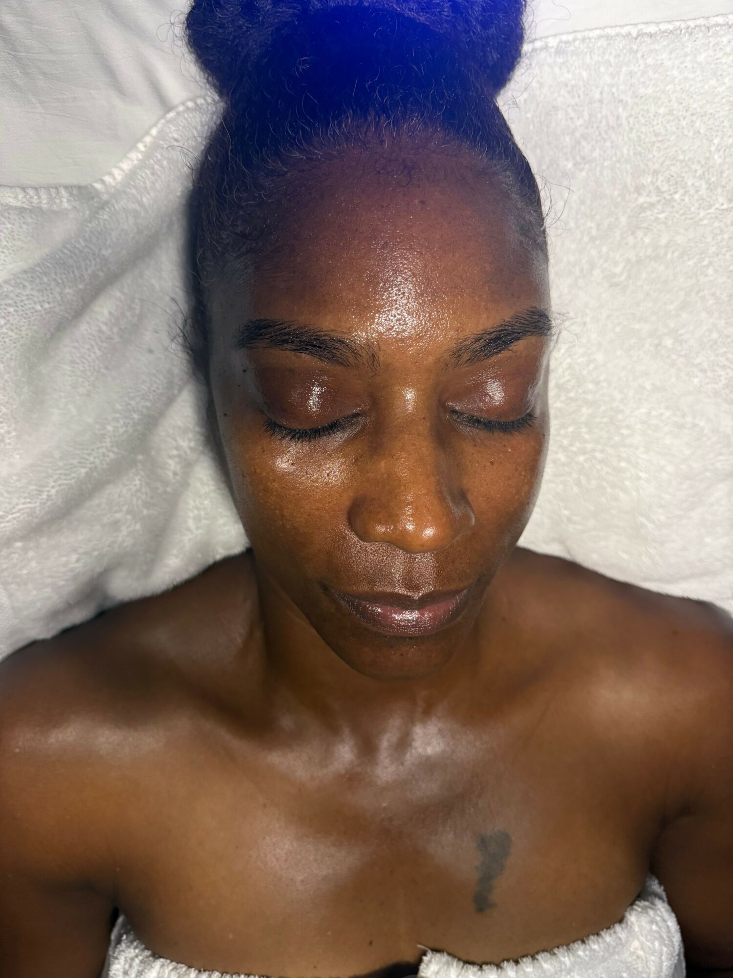 Close-up of a woman with closed eyes and a bun hairstyle, lying on a white surface, showing her shoulders and chest.