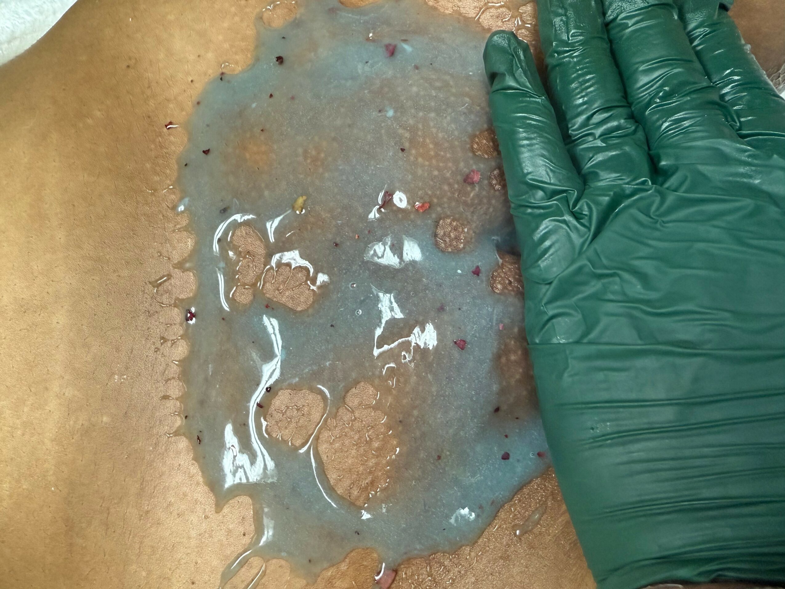Person wearing a green glove touching a wet, brown, textured surface with small spots and patches.