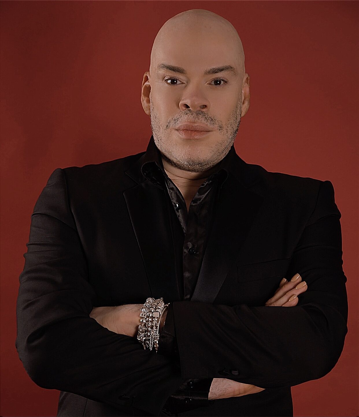 Man with crossed arms, wearing black jacket, against red background.