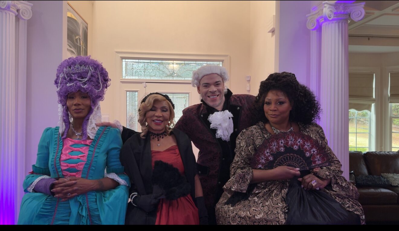 Four people dressed in colorful costumes and wigs stand indoors with columns and large windows in background.