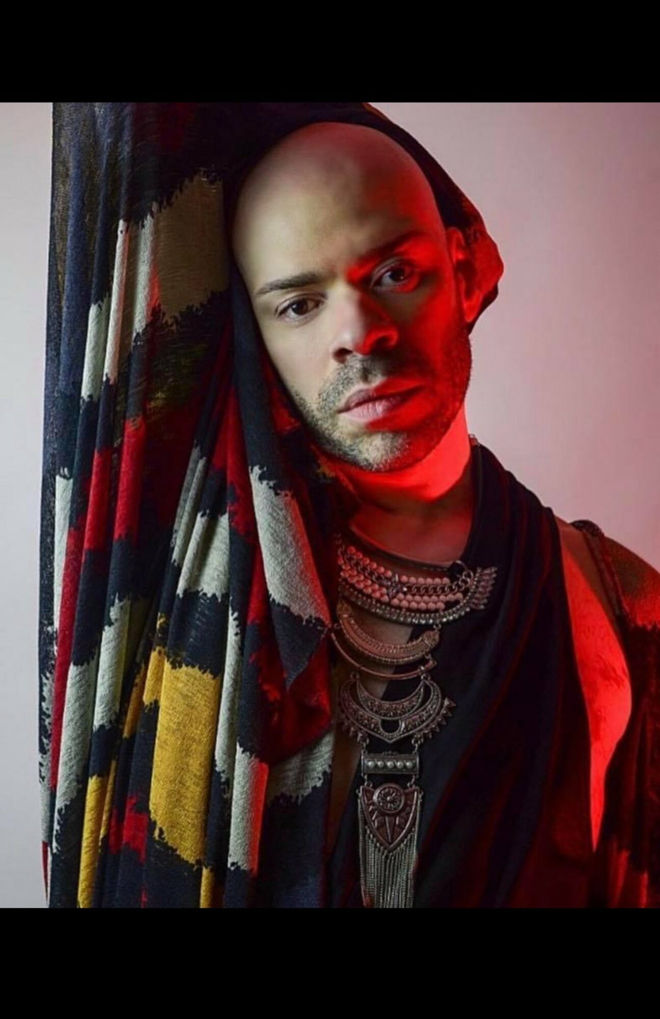Person with shaved head wearing jewelry and a colorful scarf, illuminated by red and neutral lighting.