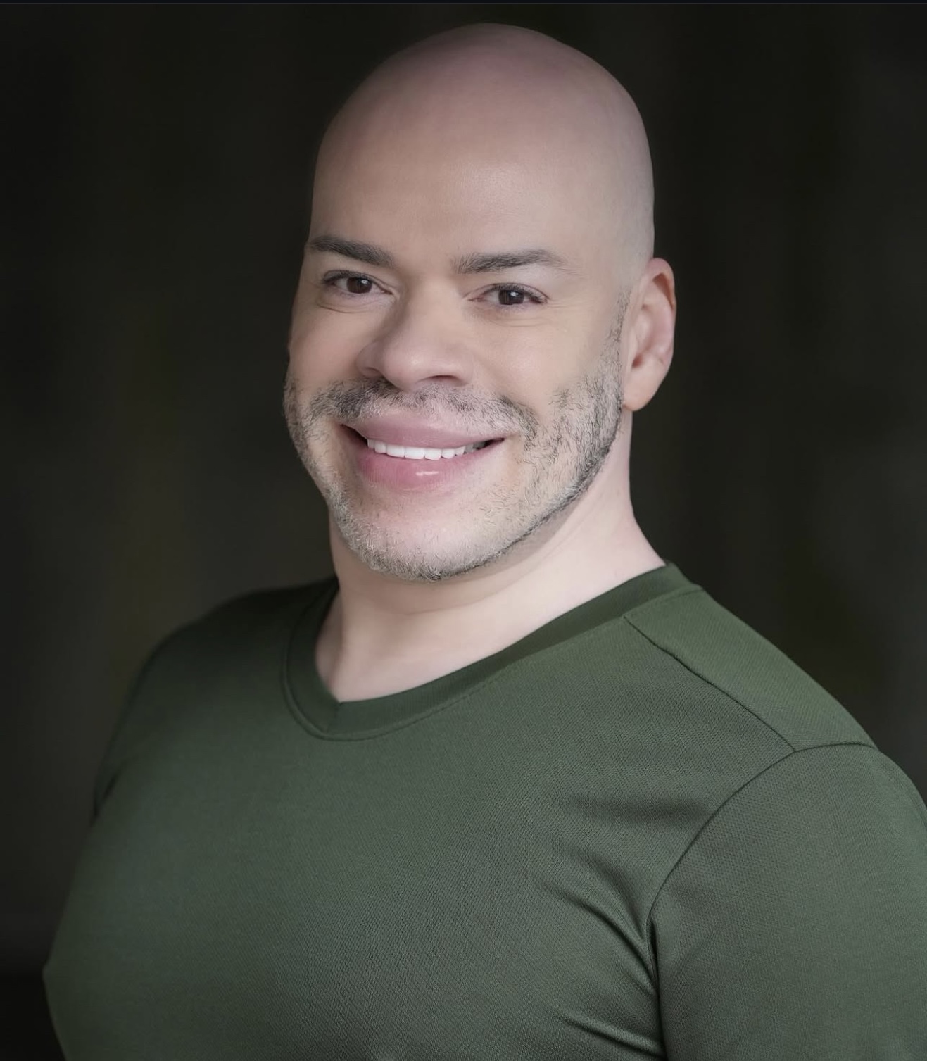 Bald man with a beard smiling, wearing a green shirt, against a dark background.