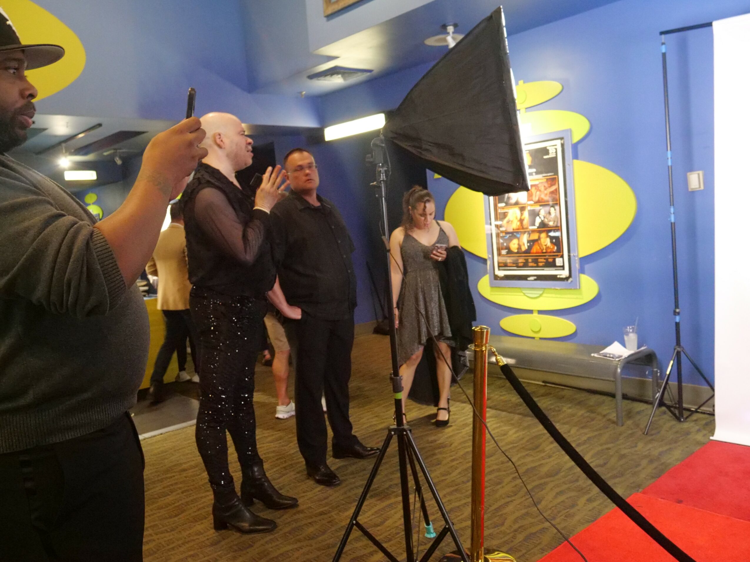 Group of people watching a screen in a colorful room with yellow and blue walls, some taking photos or videos.