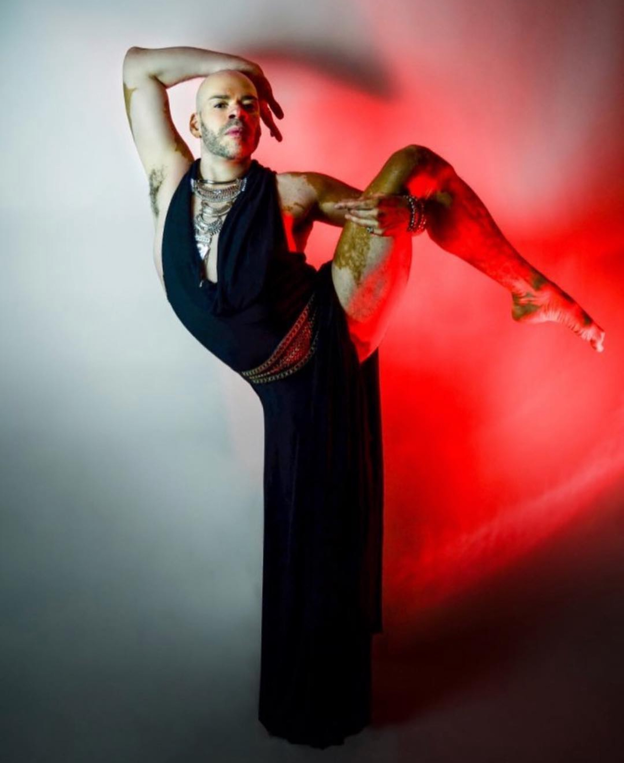 Person with tattoos in black dress poses with one leg raised and arm behind head against a gradient background.