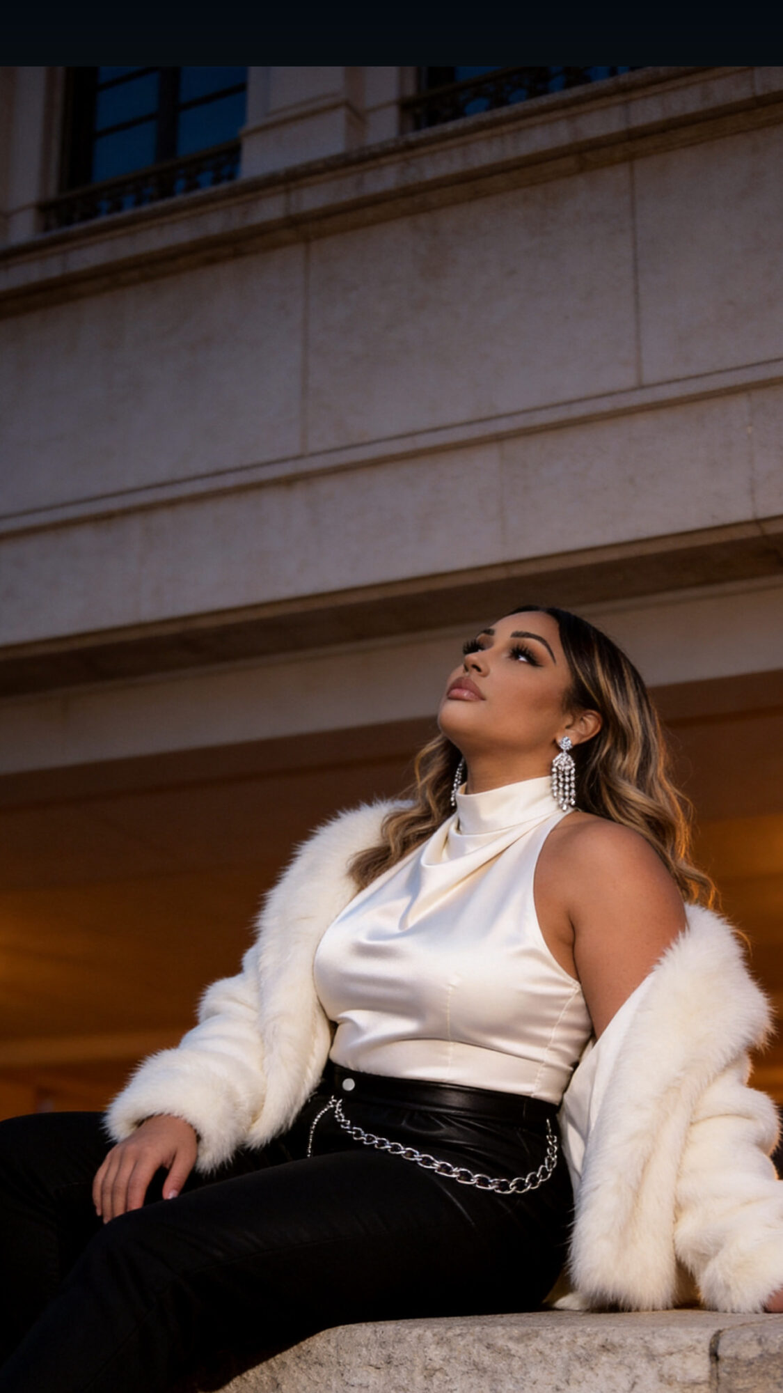 Woman sitting outdoors, looking upward, wearing a white top, black pants, and a white fur coat.