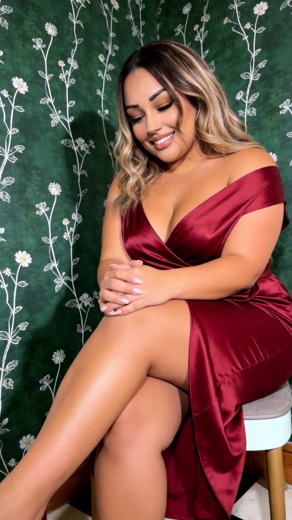 Woman in red dress sitting with crossed legs, smiling, against a green floral background.