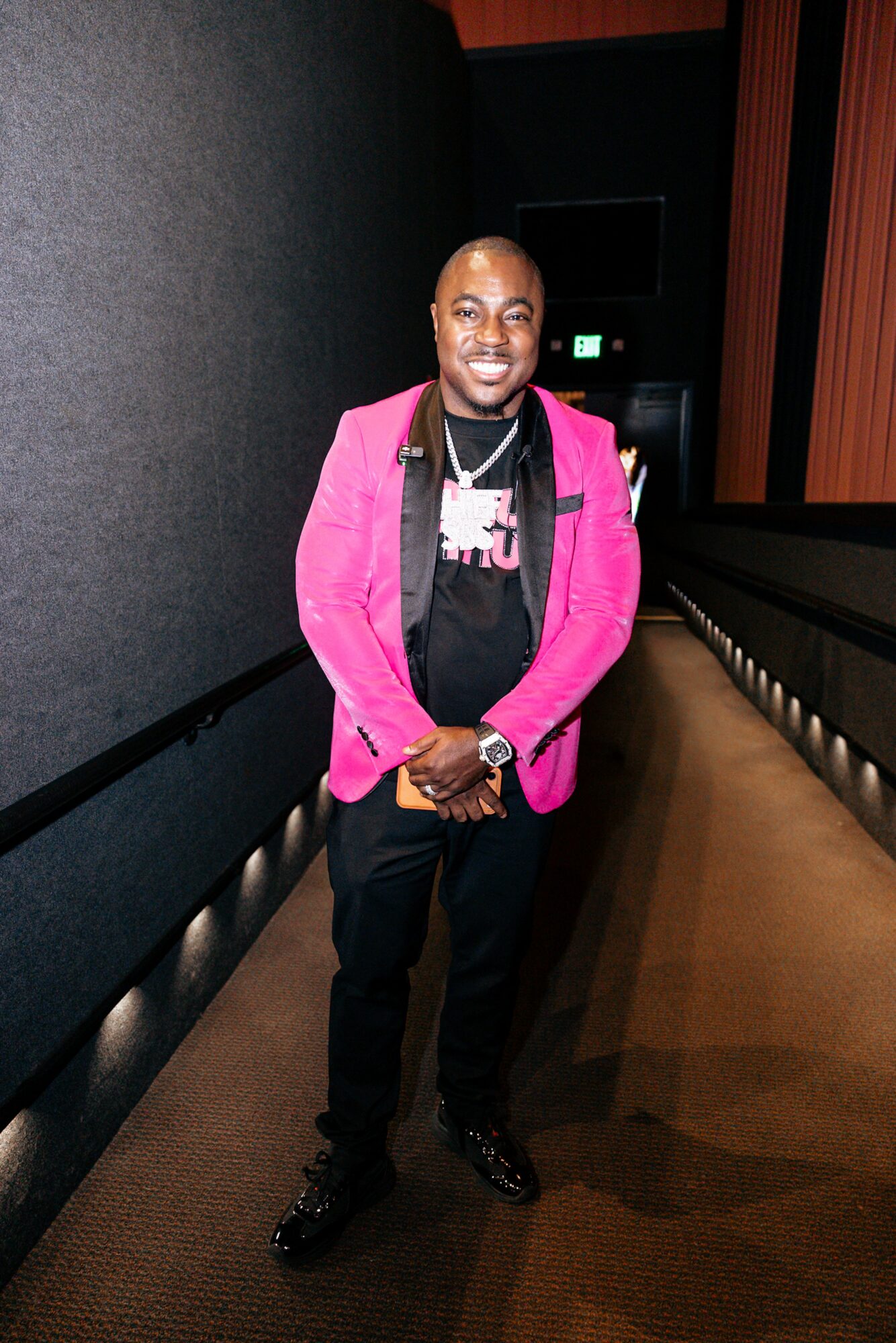 Man in pink blazer and black pants standing in a dimly lit corridor, smiling at the camera.
