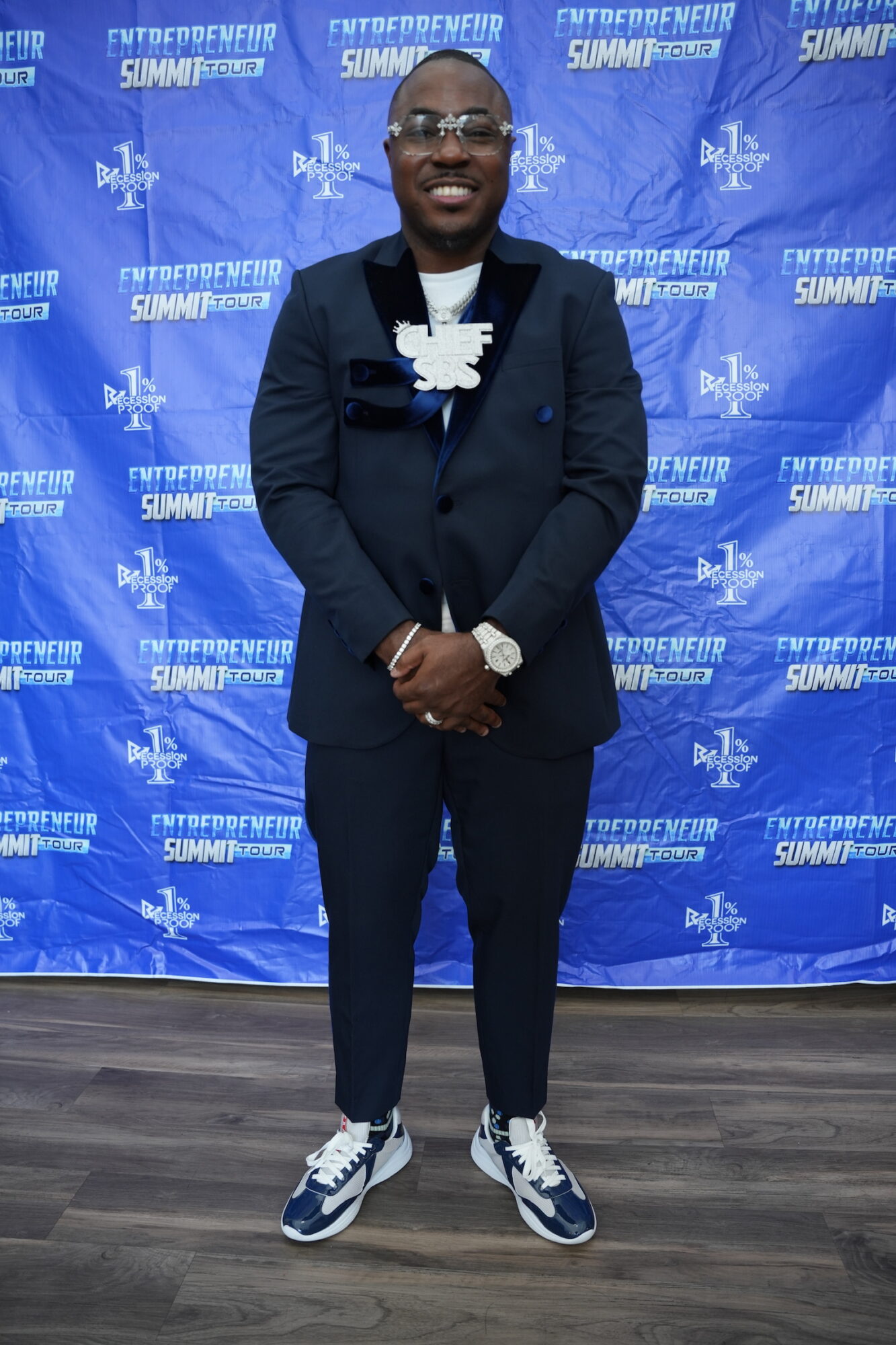 Man in a suit standing in front of a blue backdrop with event logos, smiling, wearing glasses and sneakers.
