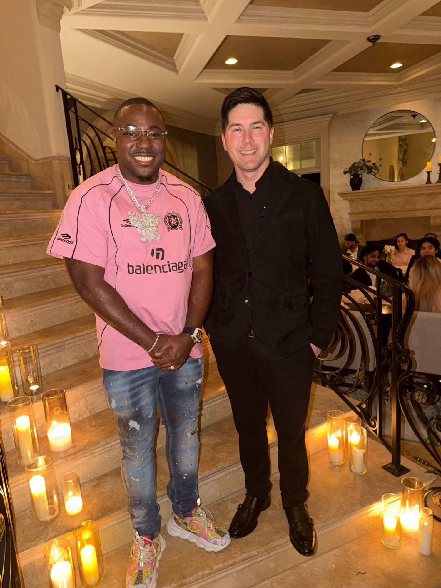Two men standing on a staircase with lit candles around, smiling at the camera in an elegant indoor setting.