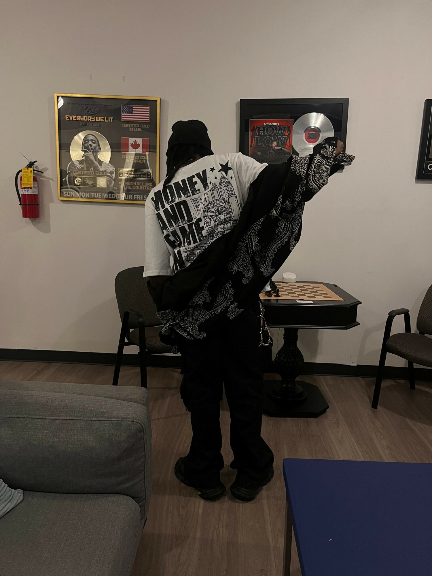 Person with a jacket and a cap, facing a wall with framed awards, reaching towards a display. Chairs and a table are nearby.