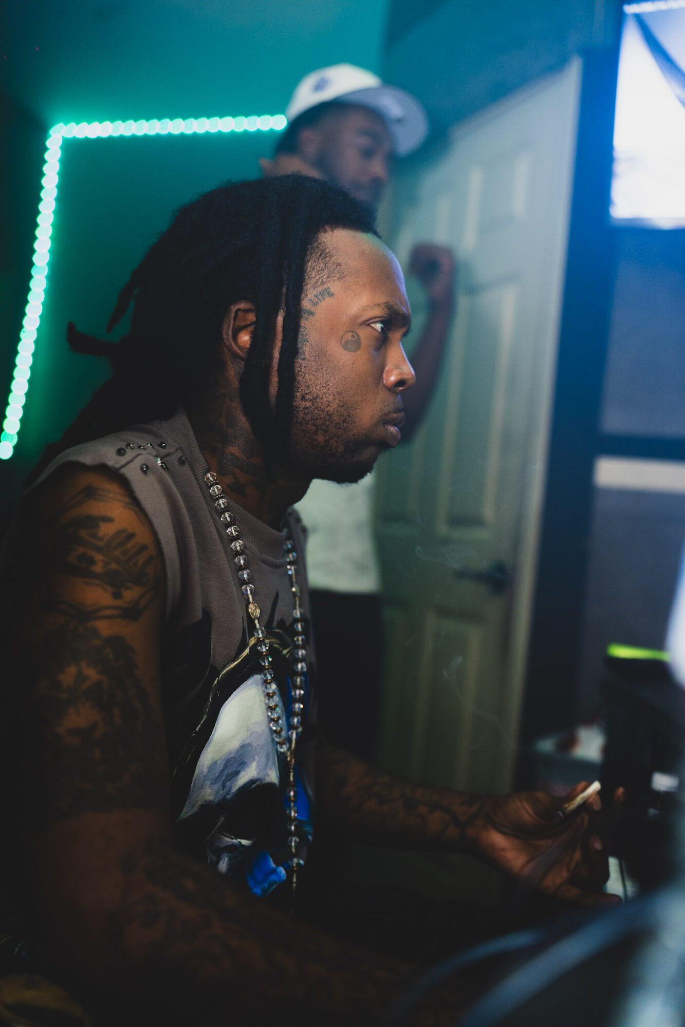 Man with dreadlocks and tattoos using a computer in a dimly lit room with neon lights.