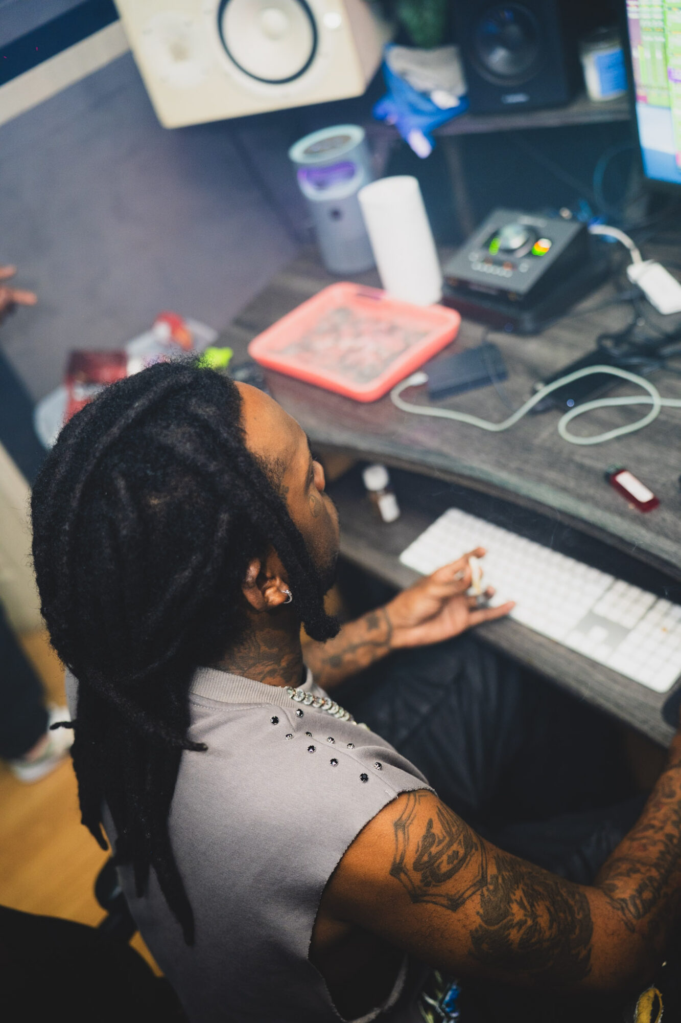 Person with dreadlocks working at a computer desk with audio equipment and supplies.