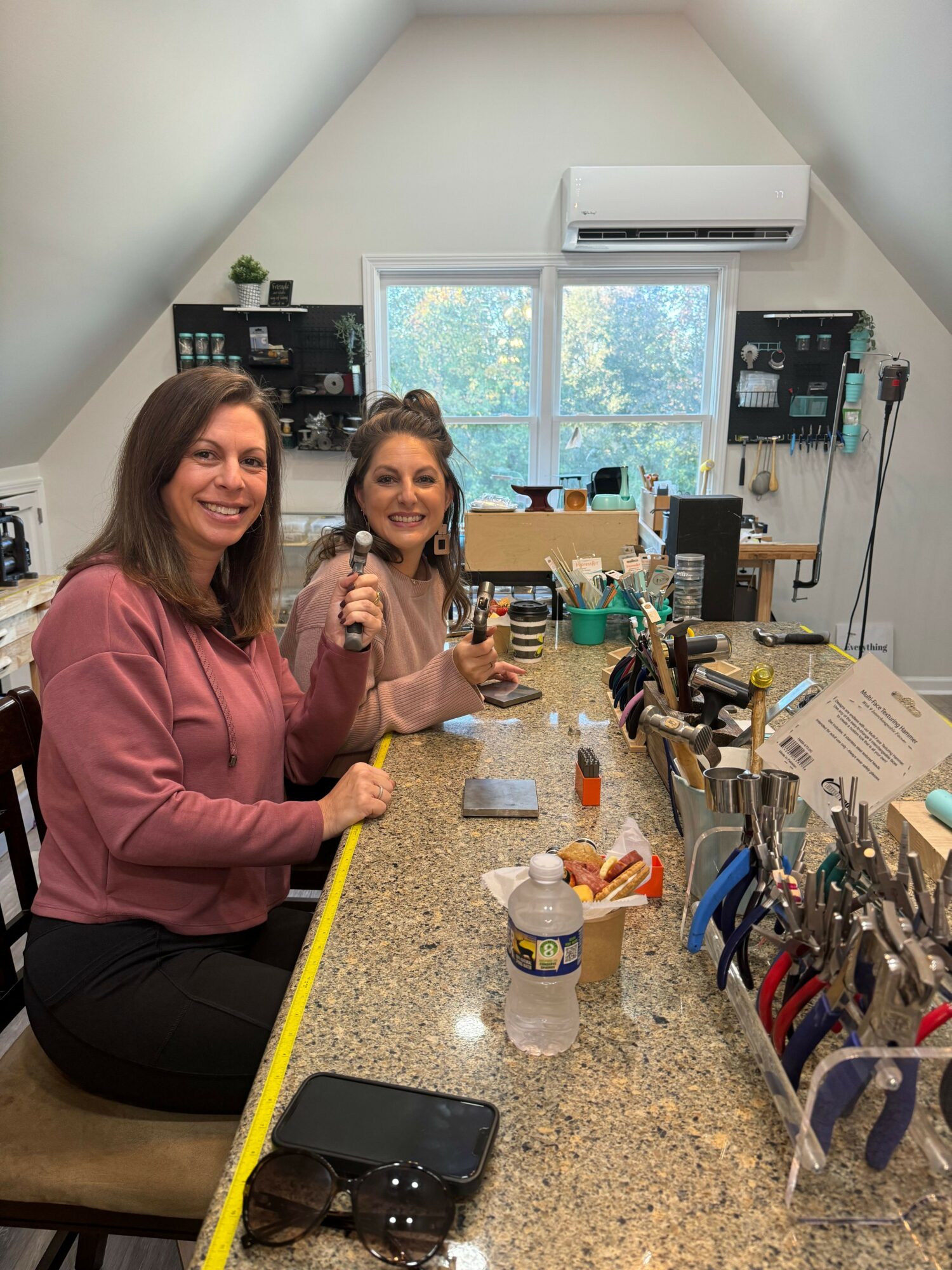 Two women sitting at a cluttered workspace with tools, bottles, and a measuring tape, smiling at the camera in a bright room.