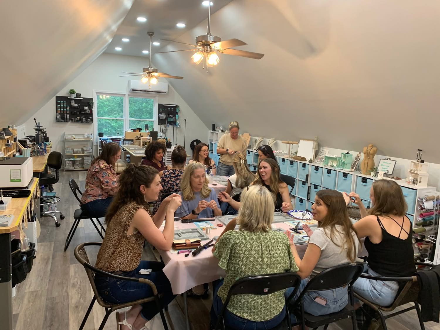 Group of women sitting around a table in a bright room with sloped ceiling, working on crafts or projects.