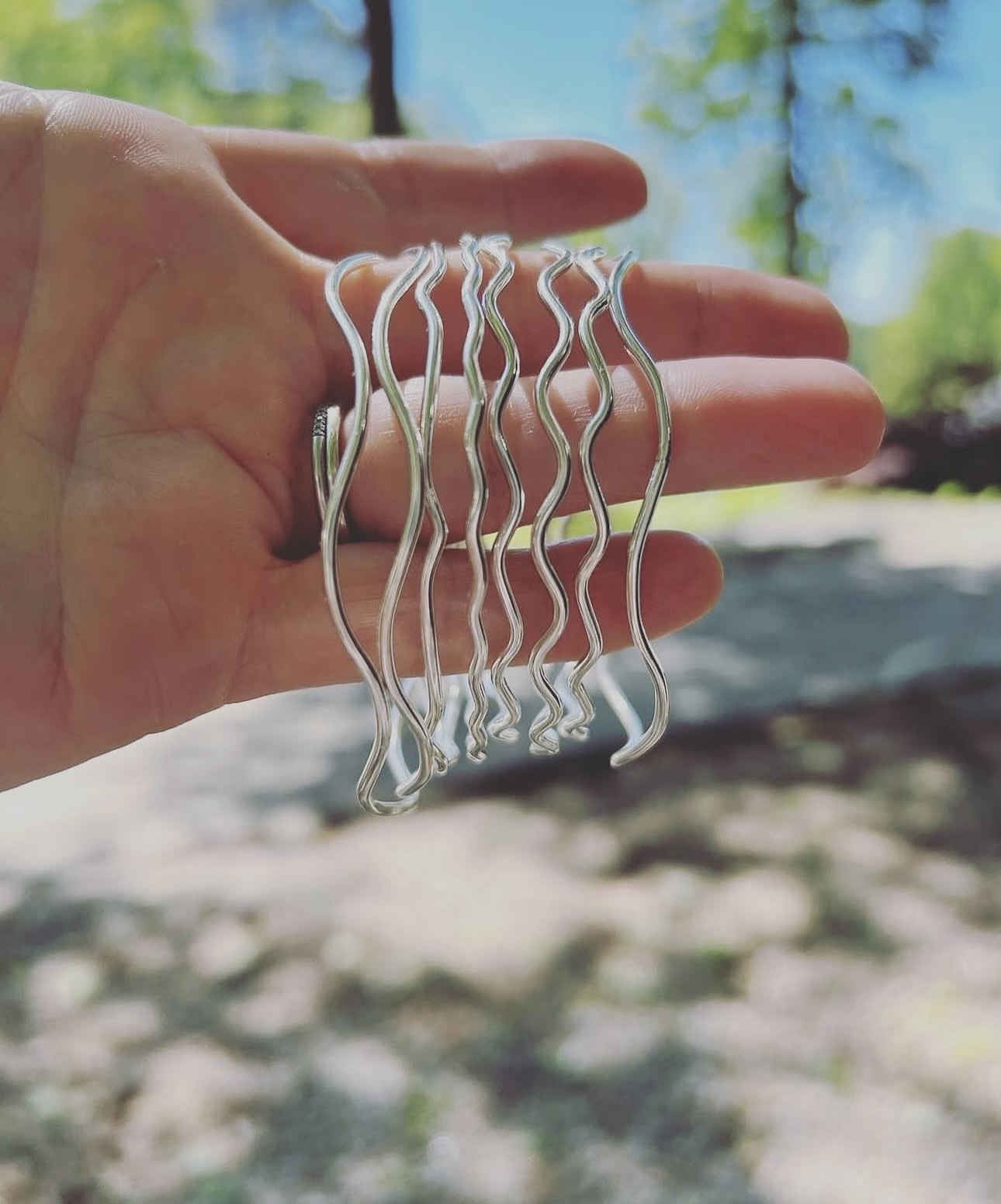 Hand holding multiple silver hair clips outdoors with trees and sky in background.