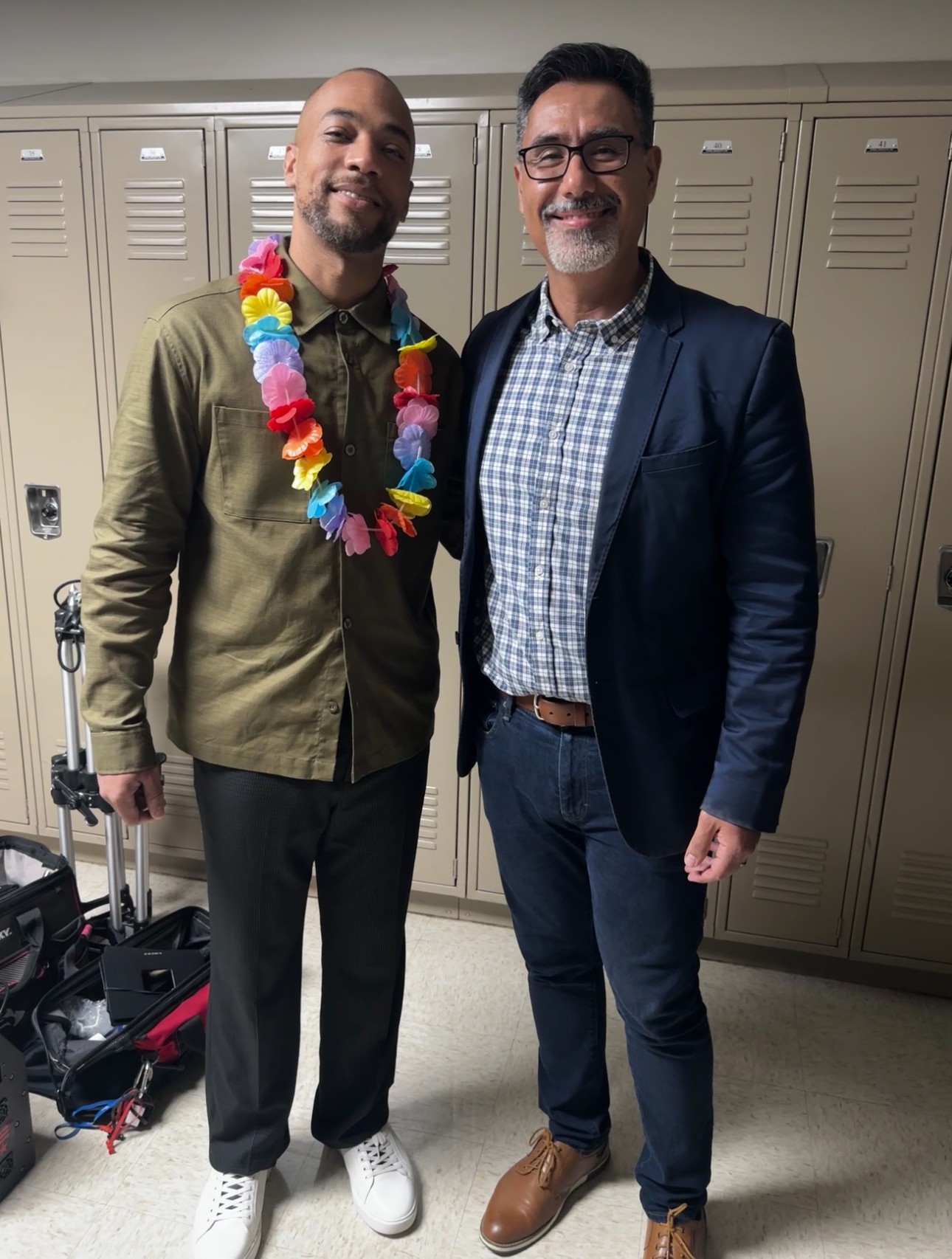 Two men standing in front of lockers, smiling, one wearing a lei, the other in a blazer and glasses.