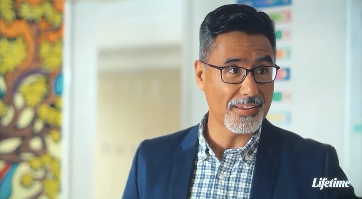 Man with glasses and a beard wearing a suit and checkered shirt, smiling indoors.