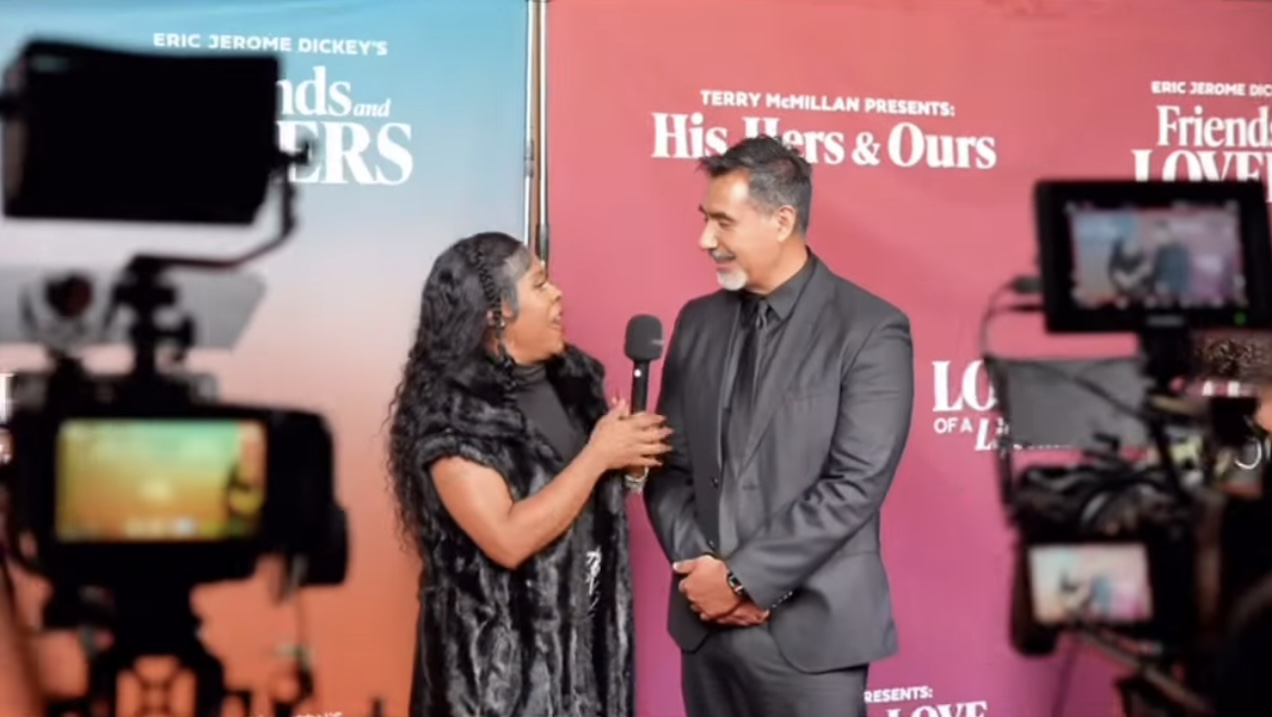 Woman interviews man on stage with pink and blue backdrop, cameras recording the event.