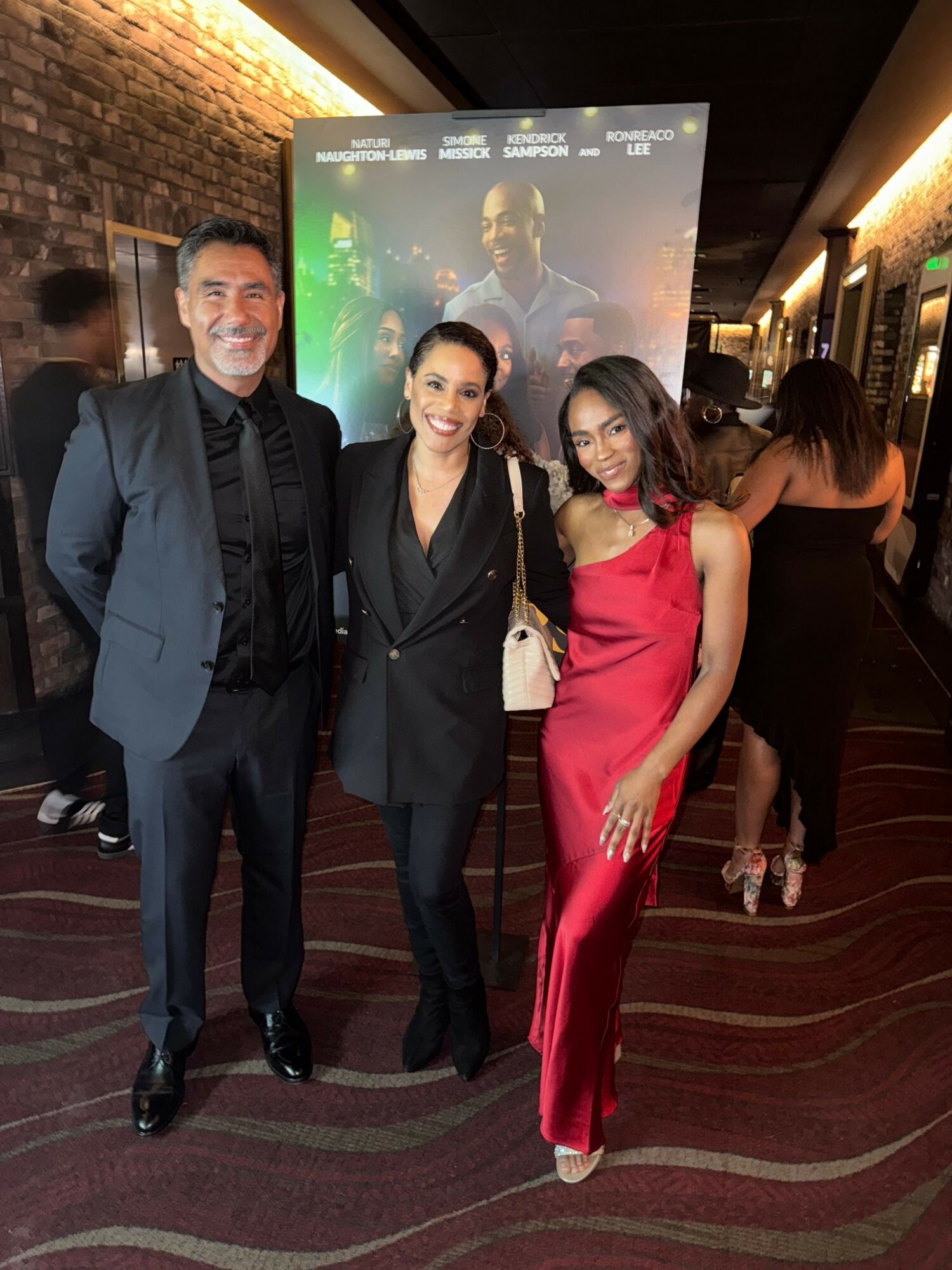 Three people in formal attire standing in front of a poster at an indoor event, smiling at the camera.