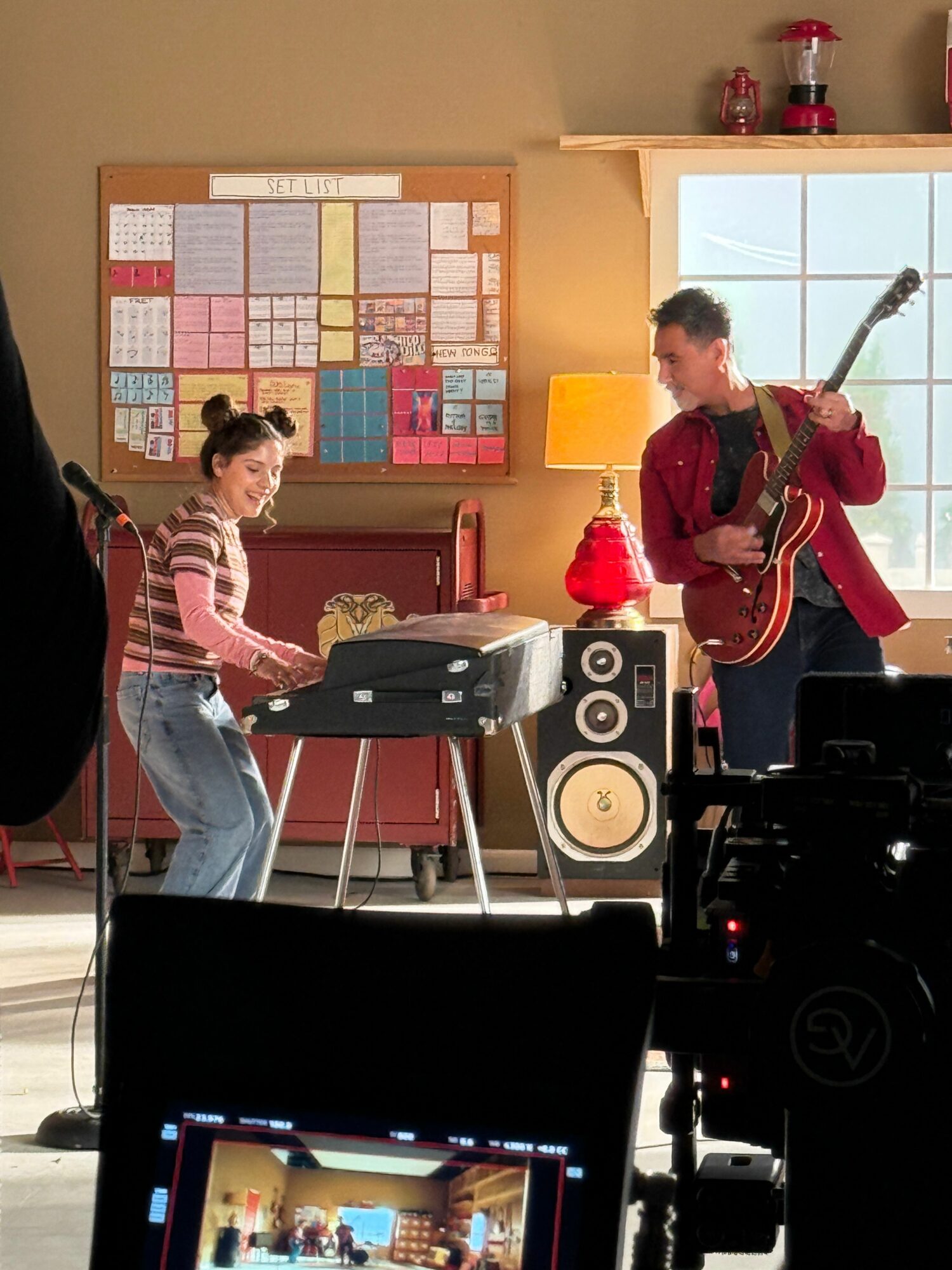 Two people in a room with musical equipment, one playing guitar and the other near a keyboard, with a bulletin board on the wall.