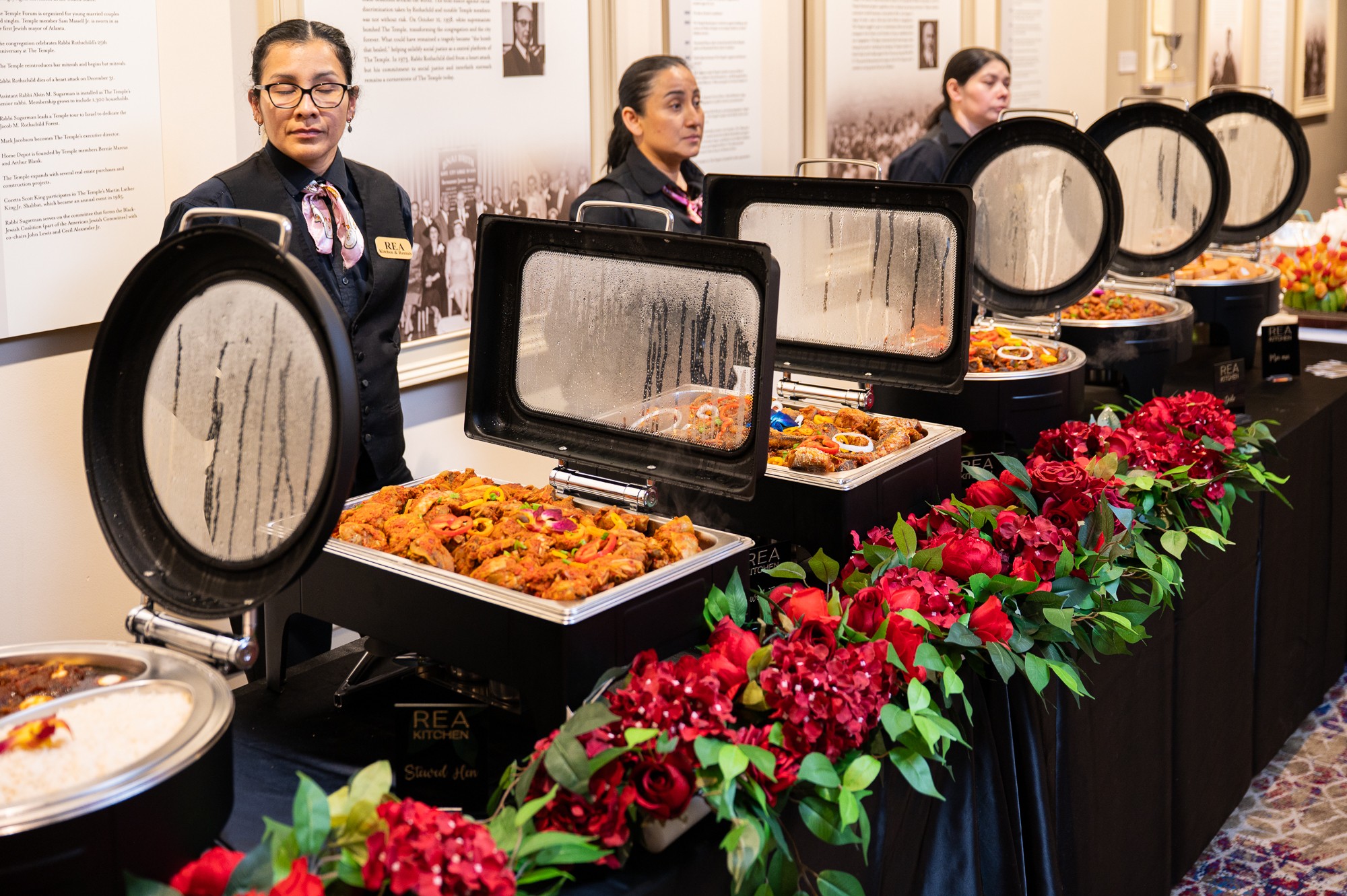 Four women in uniforms stand behind a buffet table with chafing dishes, decorated with red flowers.