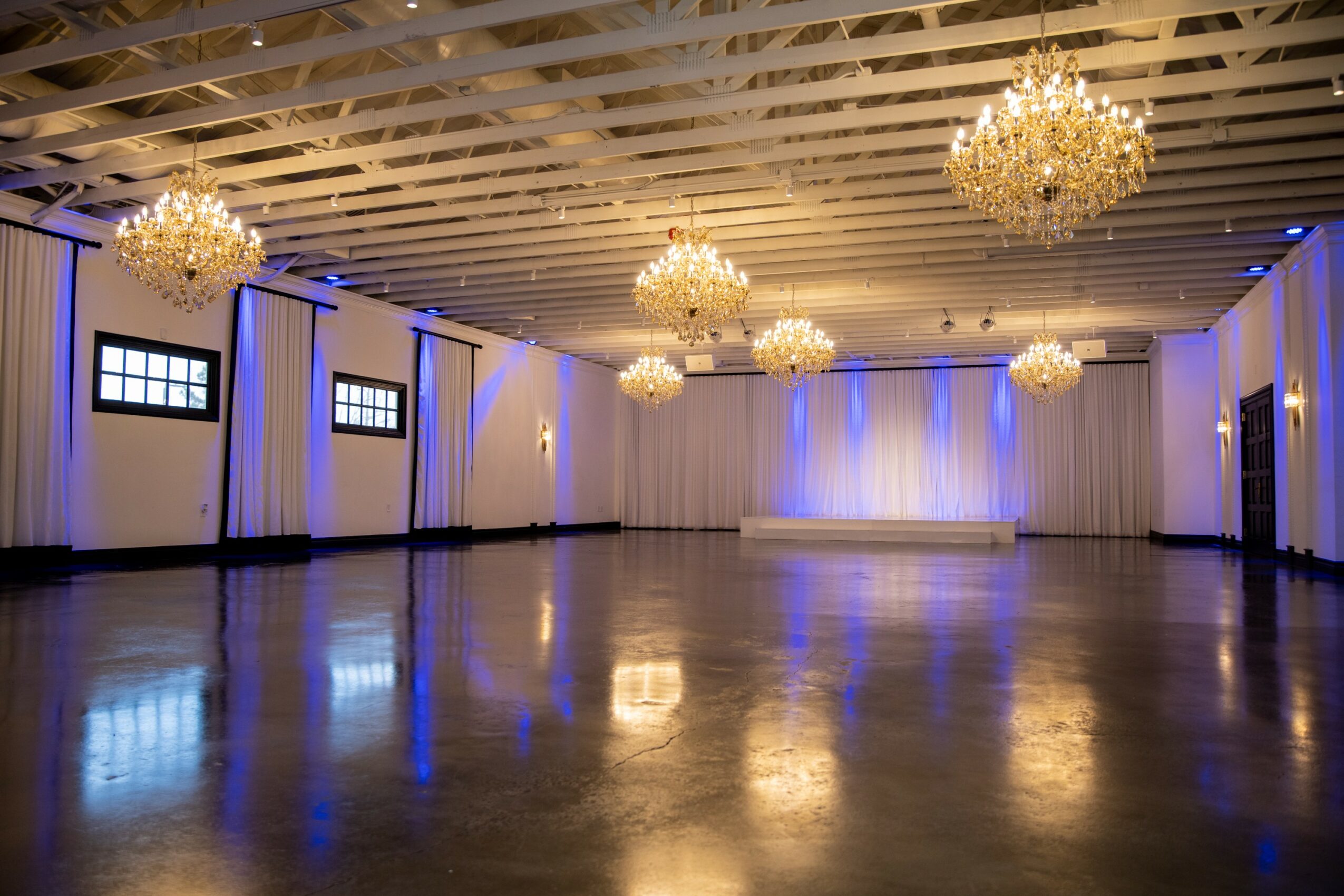 Large empty ballroom with chandeliers, white walls, and polished floor illuminated by blue lighting.