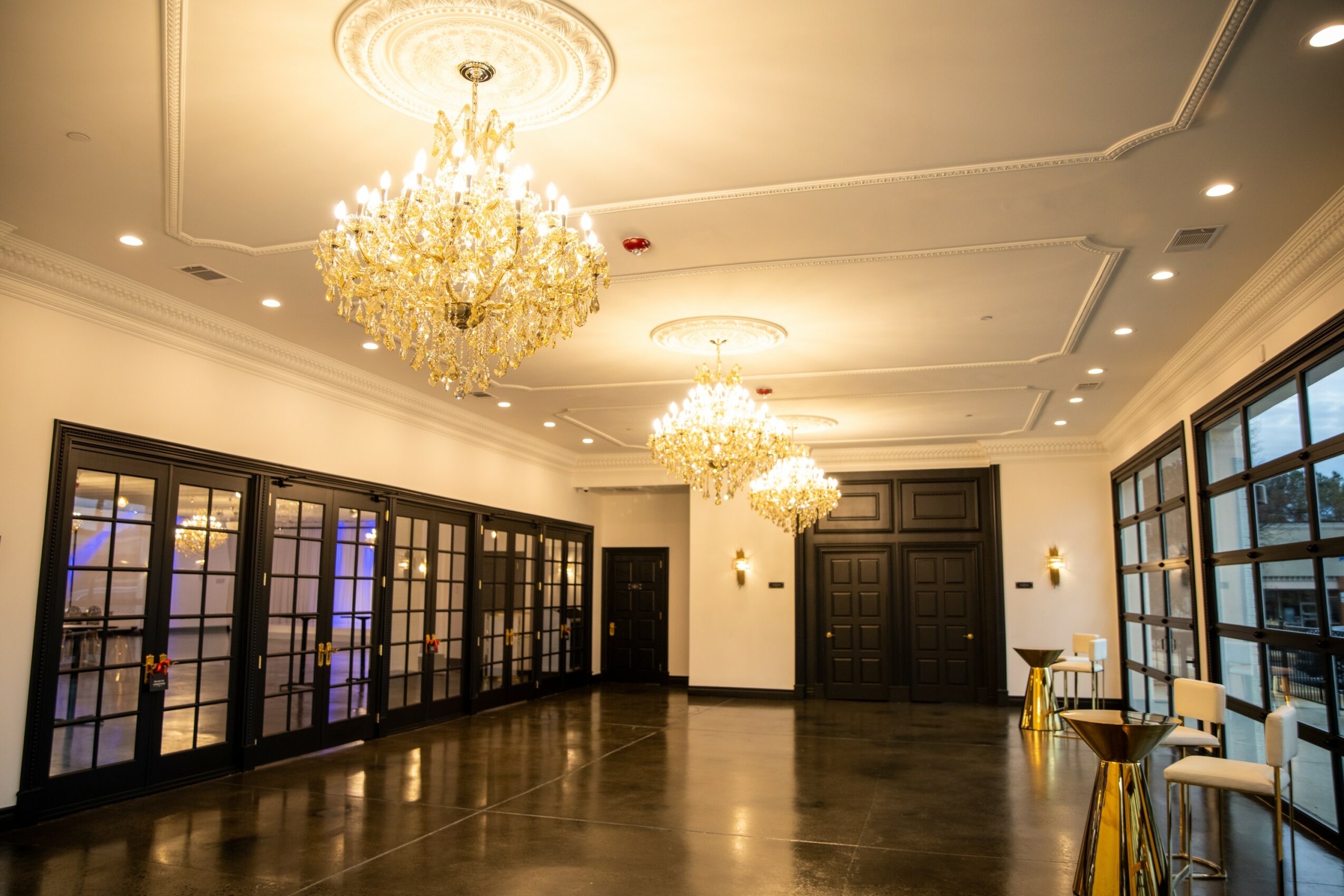 Elegant room with large chandeliers, dark doors, and glass-paneled walls, polished wooden floor, and seating area with chairs and tables.