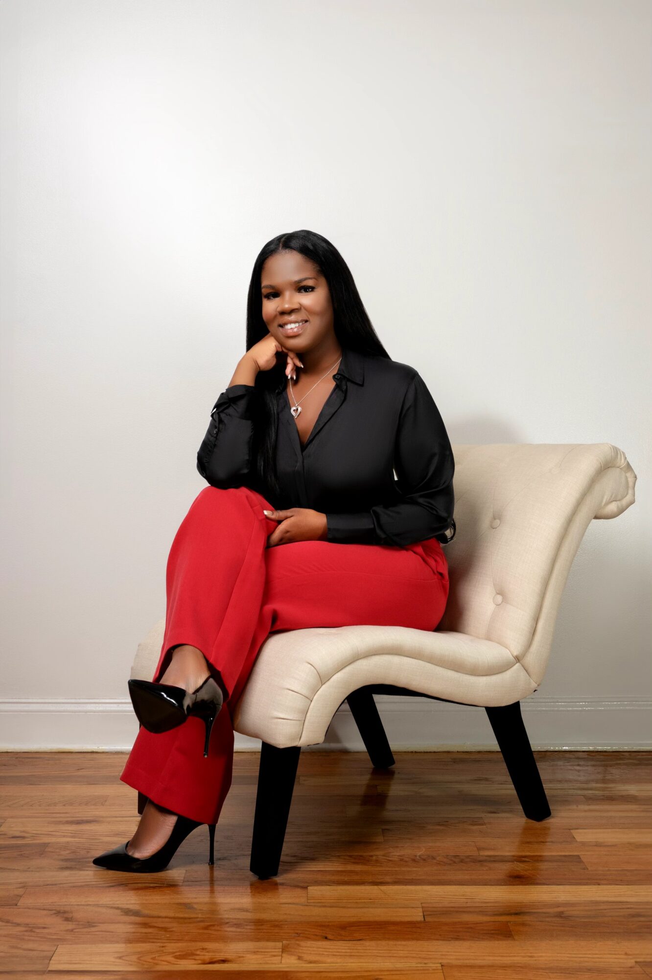 Woman sitting on a cream-colored chair, smiling, wearing a black top and red pants, with black high heels.