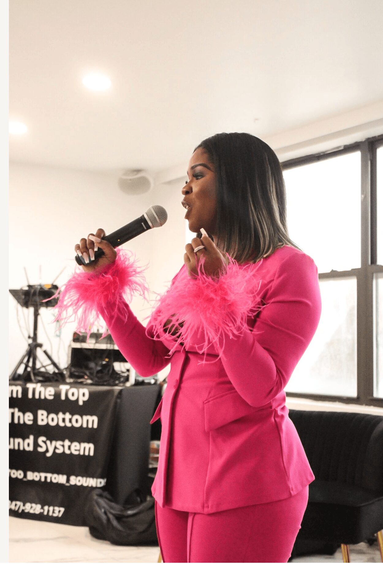Woman in pink suit with feathered sleeves holding a microphone, speaking indoors near a window.
