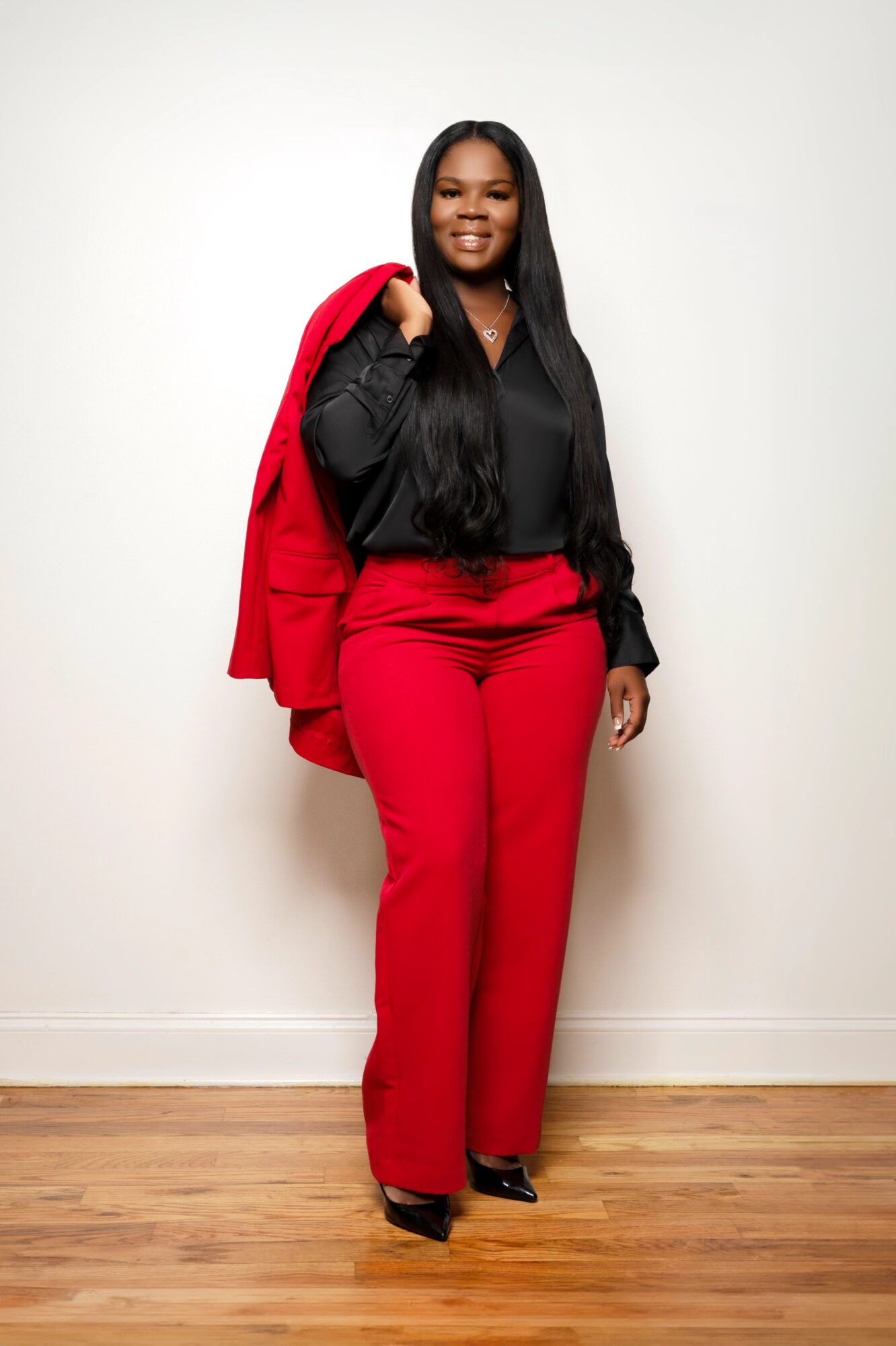 Woman standing against a white wall, wearing black top and red pants, holding a red jacket over her shoulder.