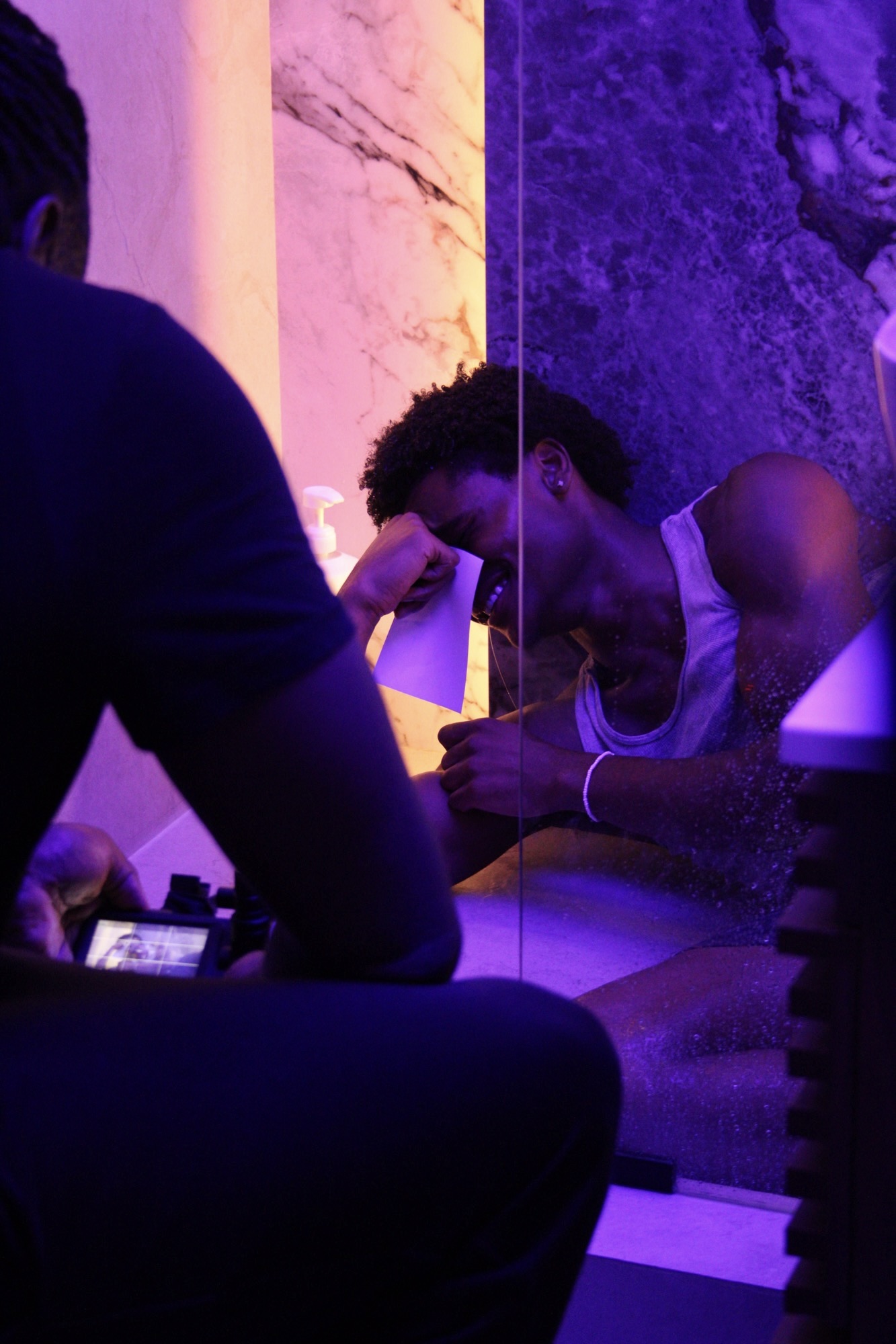 Person with curly hair and tank top looking into mirror, hand on forehead, in dim purple-lit room.