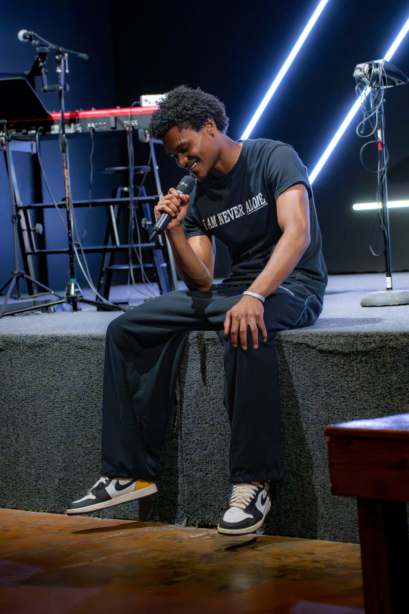 Young man sitting on stage holding a microphone, smiling, with musical equipment and bright lights behind him.