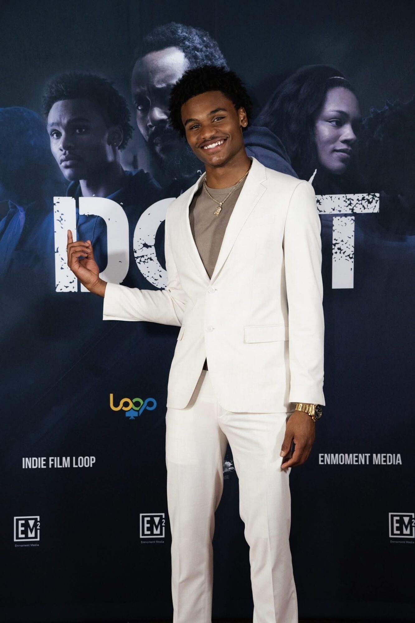Young man in white suit smiling, holding a card, standing in front of a dark poster with faces and text.