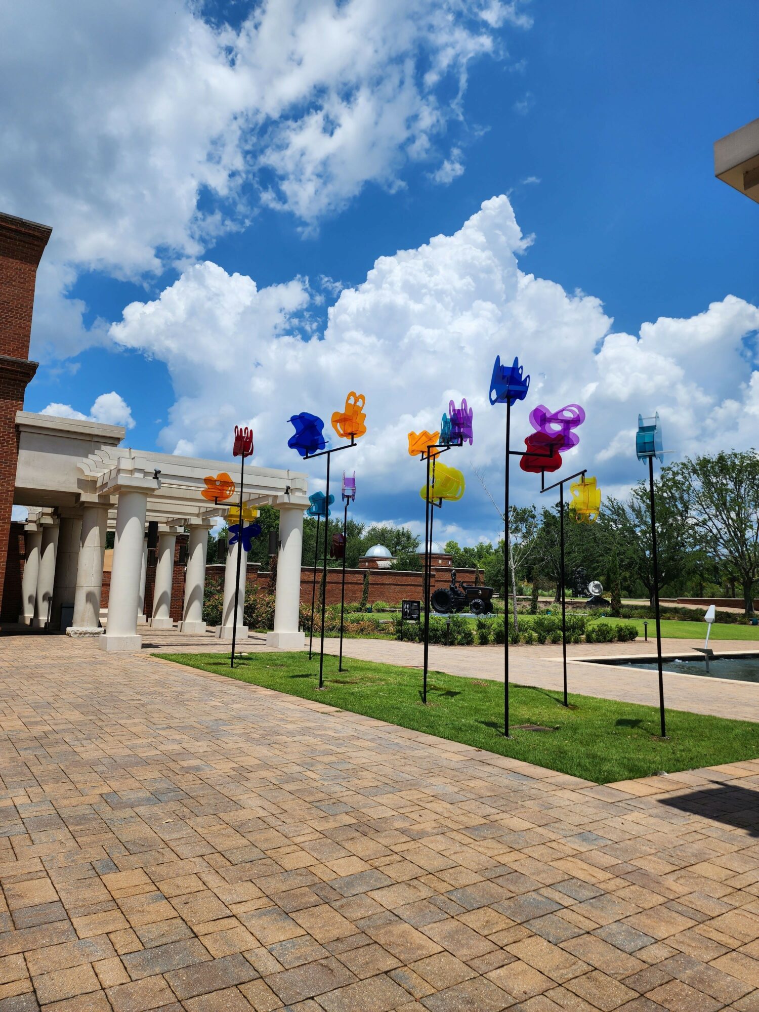 Colorful wind sculptures on poles in a grassy area with a building and trees, under a partly cloudy sky.