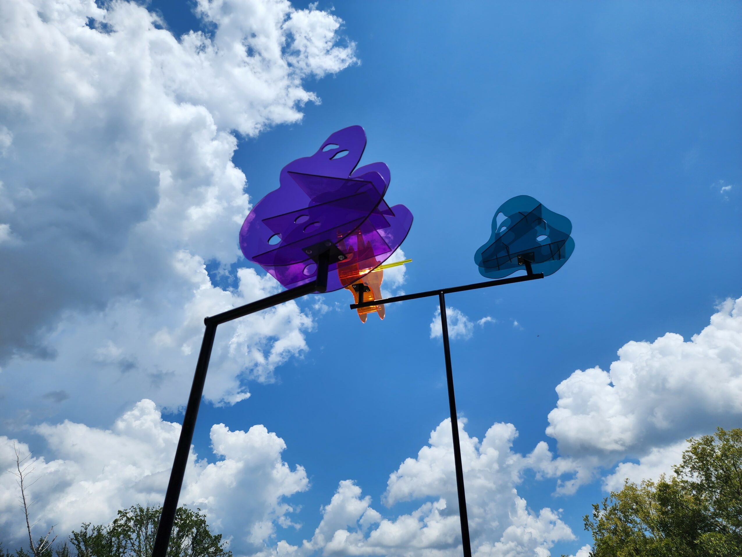 Colorful pinwheels spinning on a pole against a blue sky with clouds and trees in the background.