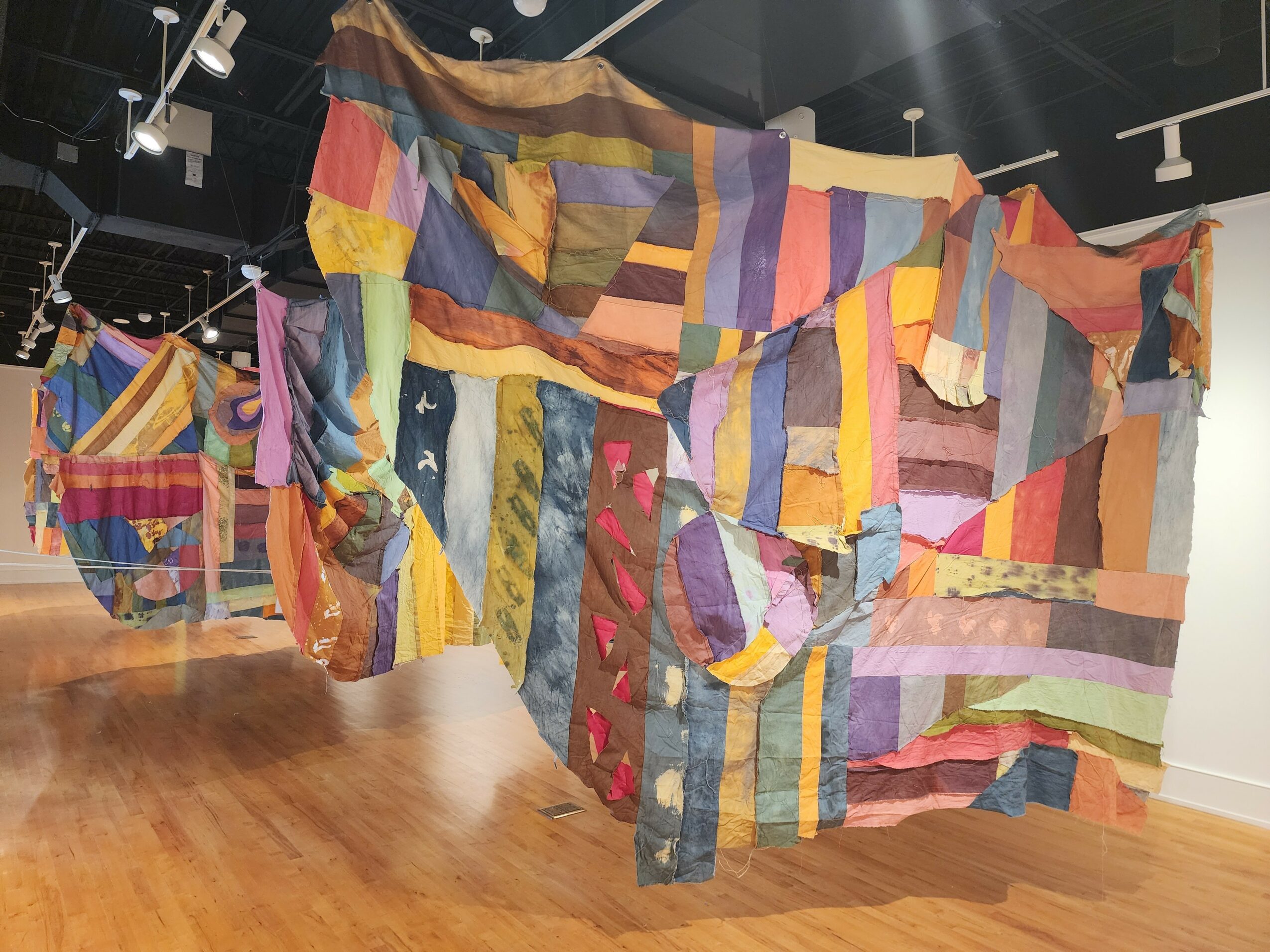 Large colorful fabric installation with various patterns and textures hangs in an indoor gallery space.