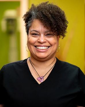 Smiling woman with curly hair, wearing earrings and a necklace, in front of a yellow background.