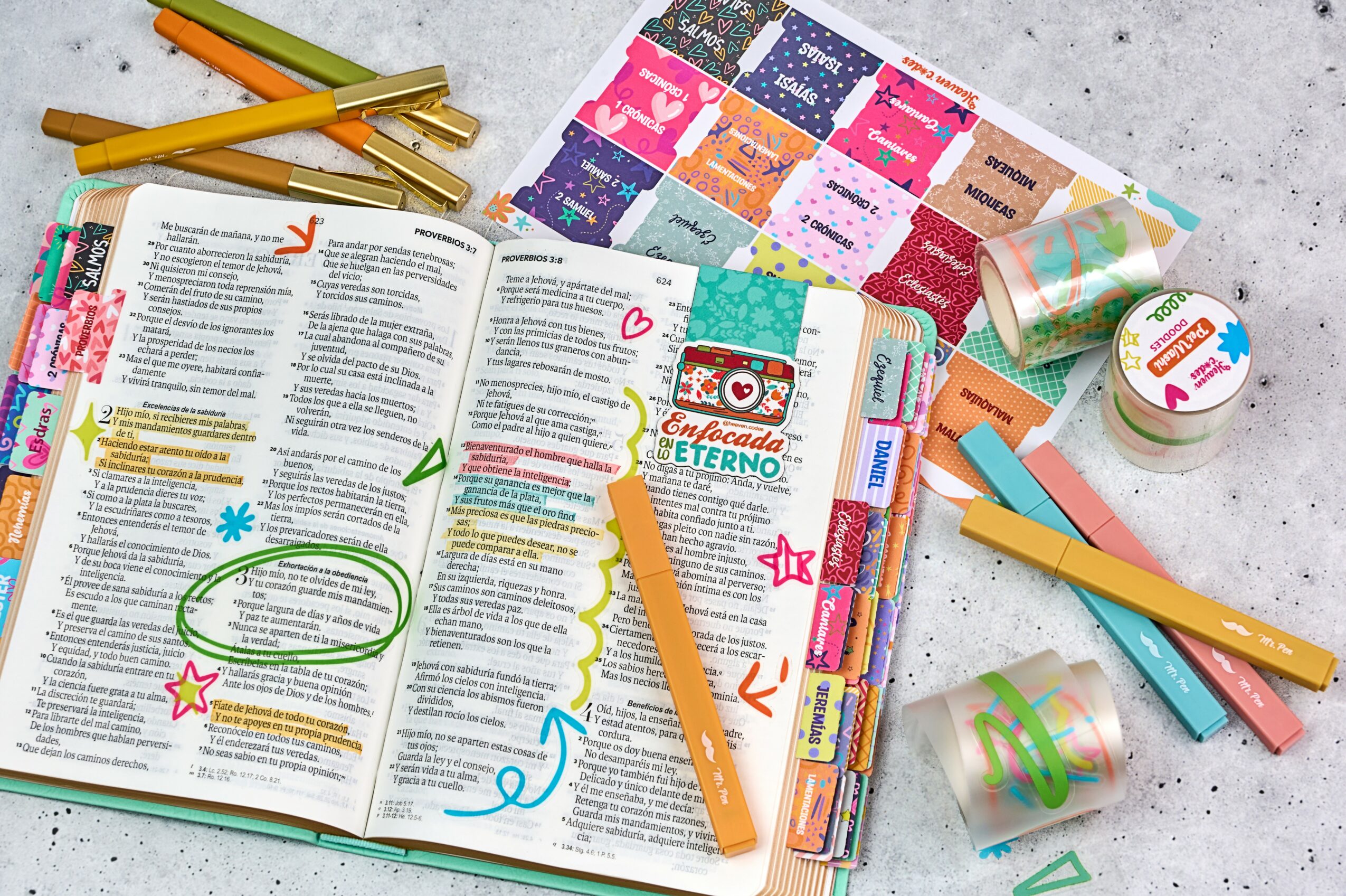 Open book with colorful pens, sticky notes, and stationery on a gray surface.