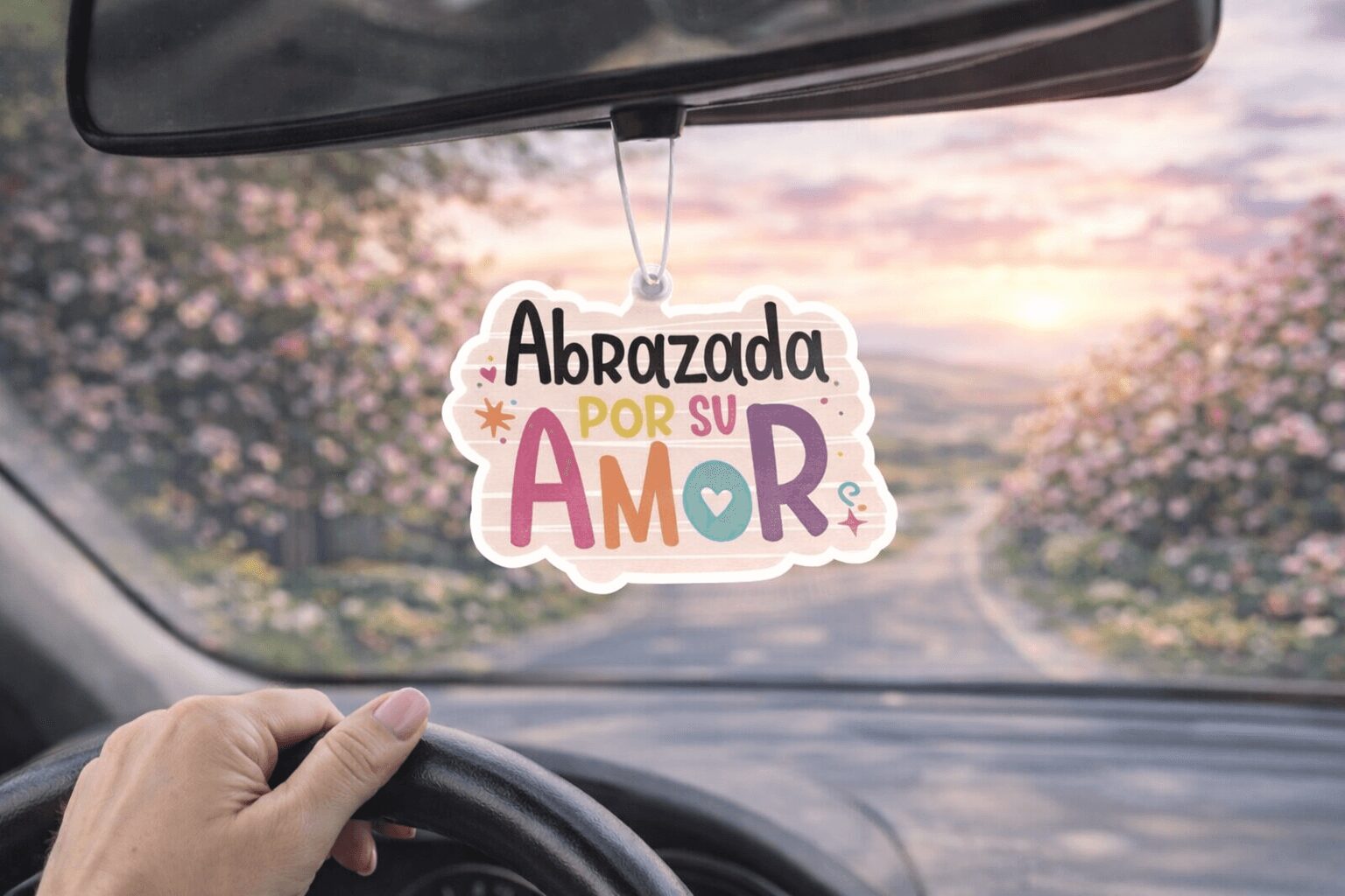 Car interior with a hand on the steering wheel and a decorative air freshener hanging from the rearview mirror, blurry background.