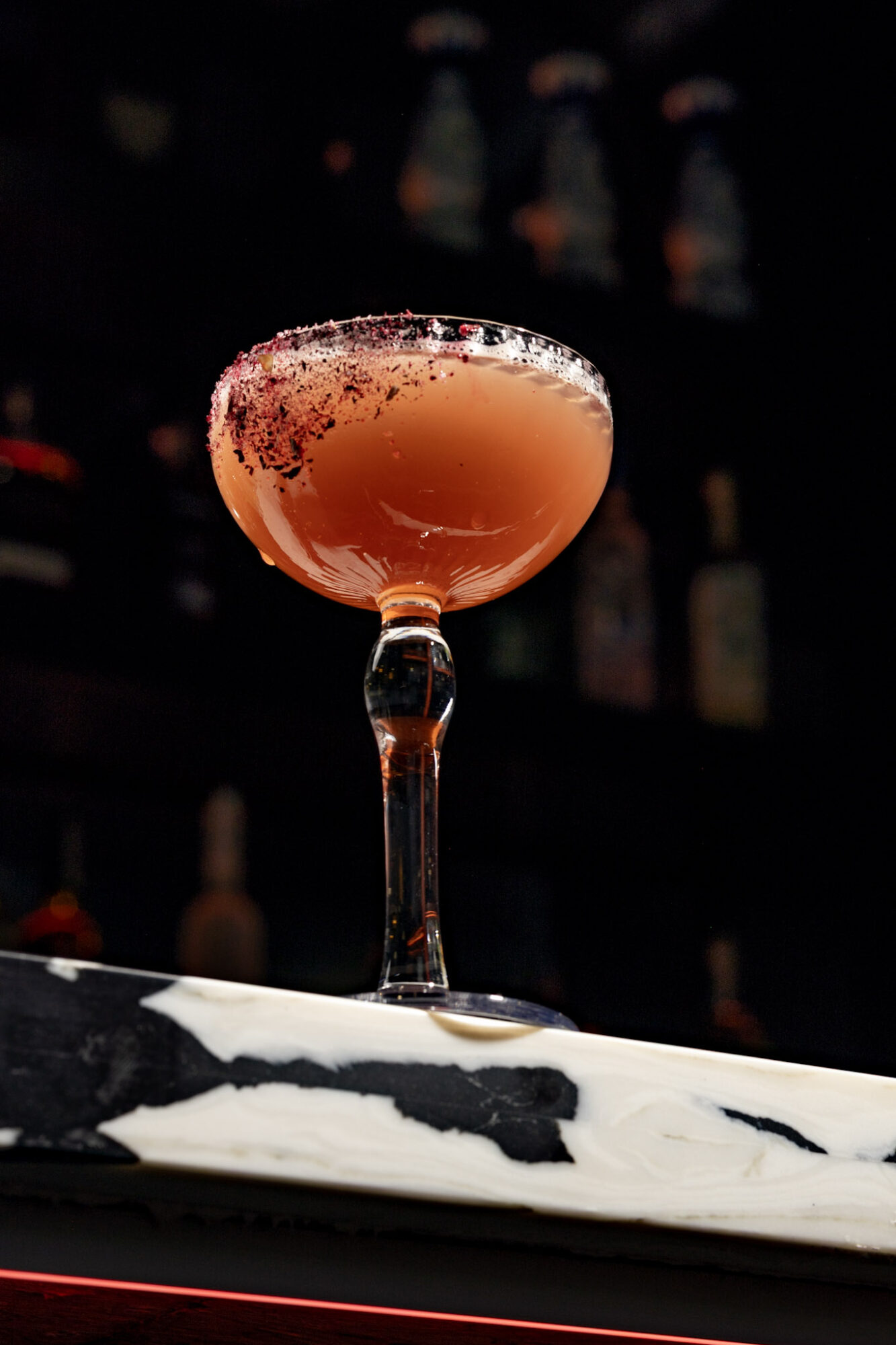 Cocktail glass with pink drink and frothy top on a marble surface, dark bar background with bottles.