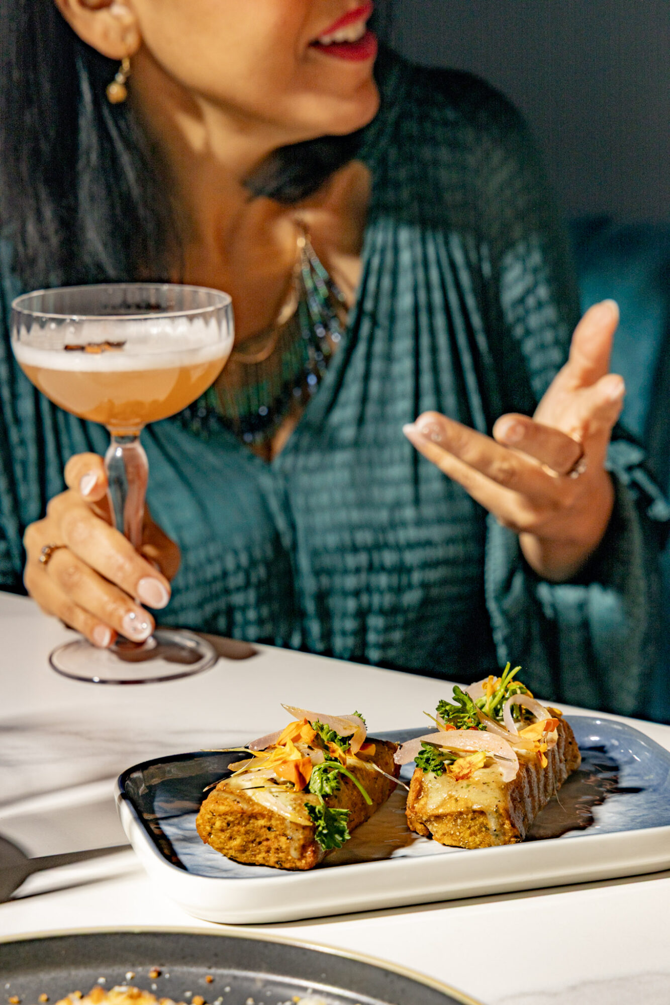 Woman holding a cocktail glass with food on a plate in front, wearing a teal top and jewelry.