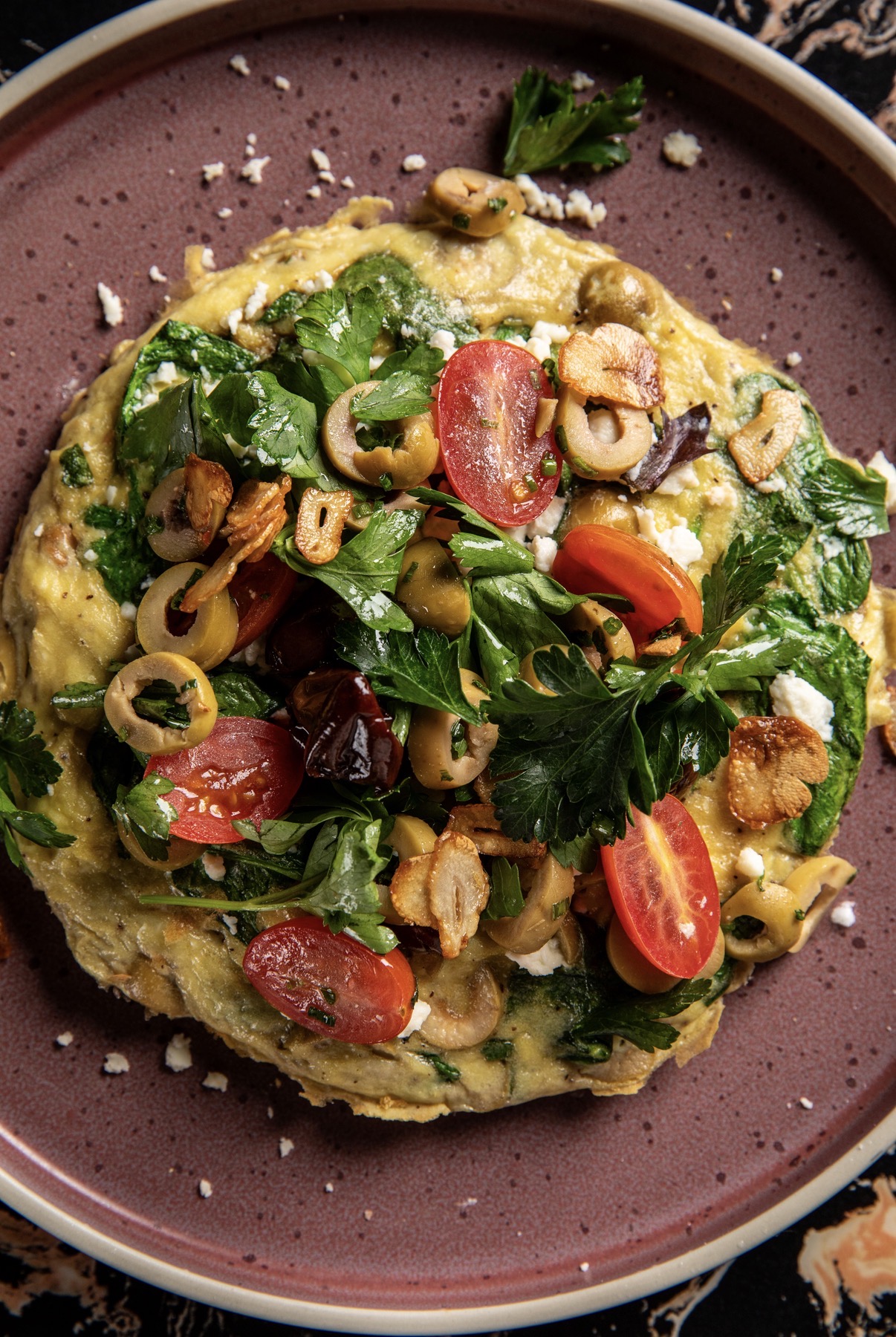 Vegetable omelette topped with cherry tomatoes, spinach, olives, and herbs on a purple plate.