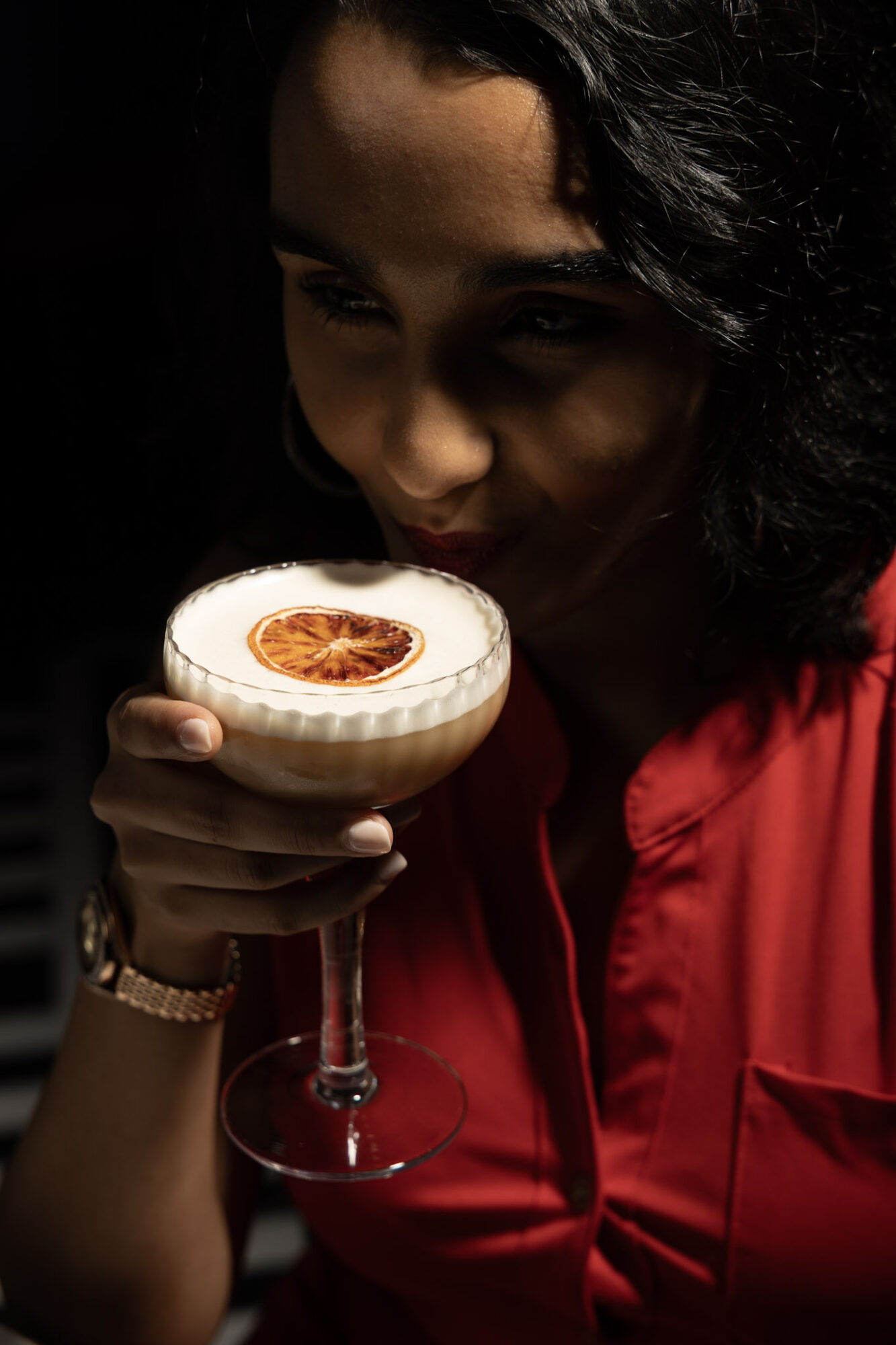 Woman holding a cocktail glass with a dried citrus slice garnish, wearing a red top, in a dark setting.
