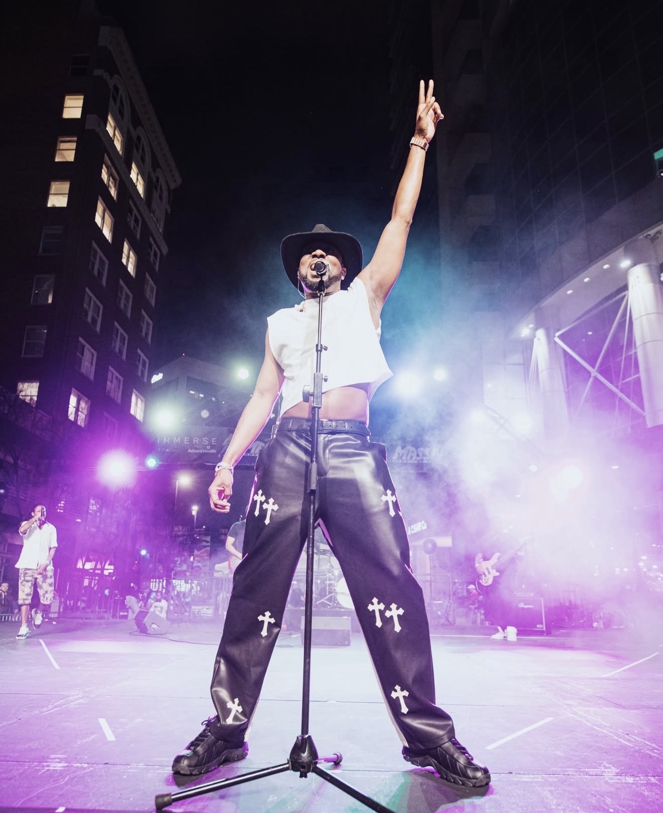 Performer on stage with one arm raised, wearing a hat, sunglasses, and baggy pants with crosses, in front of city buildings.