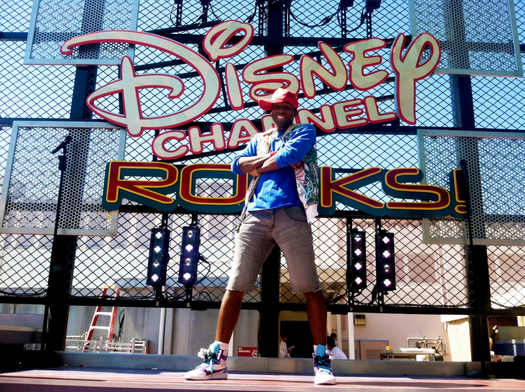Person standing on stage with arms crossed in front of a Disney Channel Rocks sign and a chain-link fence background.