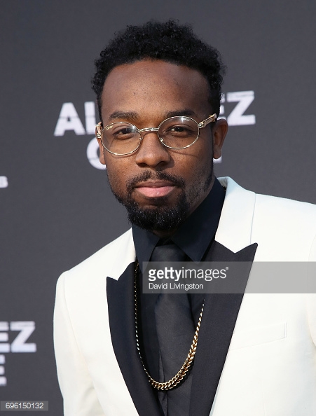Man with glasses, beard, wearing a tuxedo with a white jacket and black shirt, standing against a dark background.